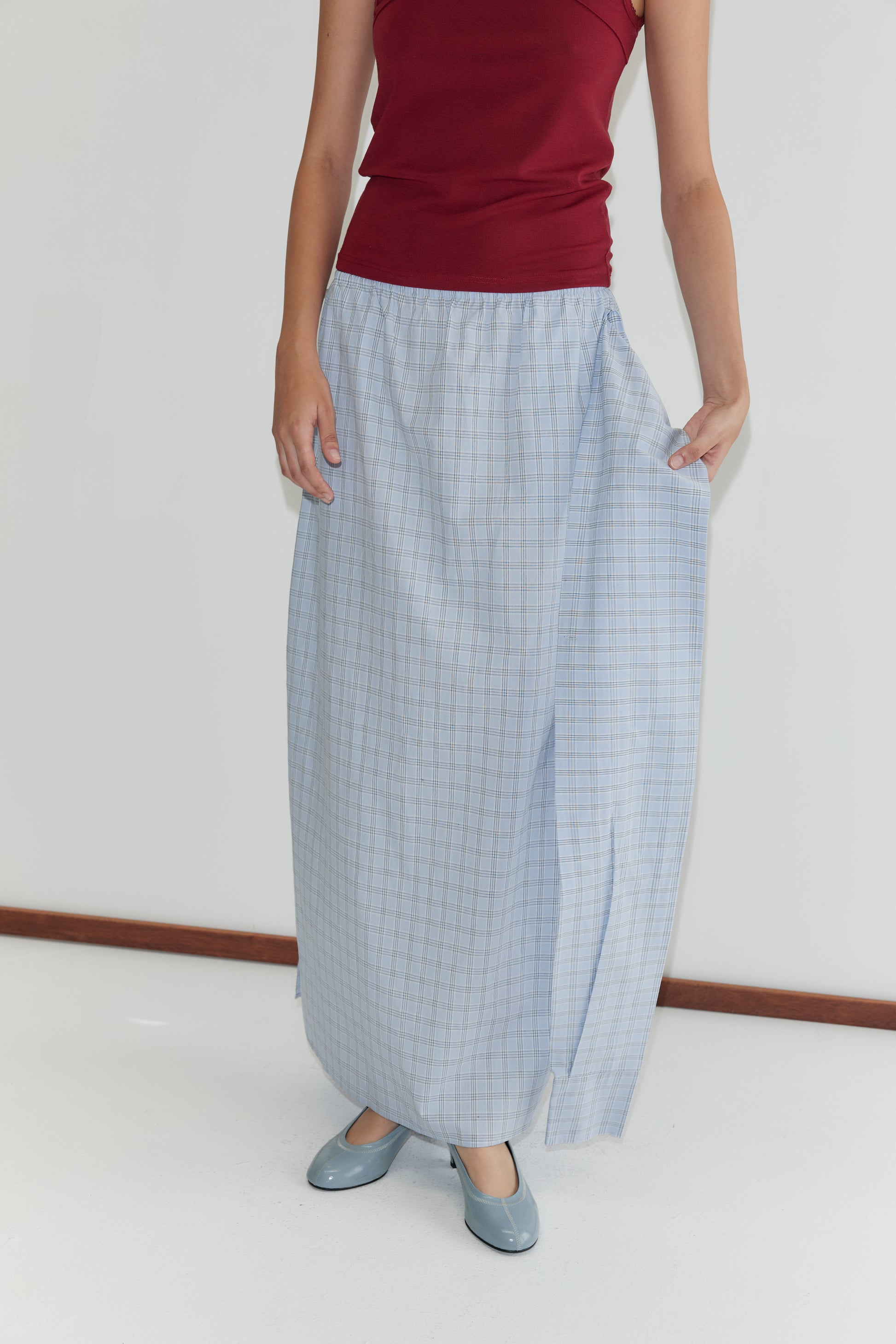A person wearing a sleeveless red top, light blue shoes, and the Way Skirt - Powder Check from Deiji Studios stands against a plain white background, holding the side of the full-length plaid skirt.