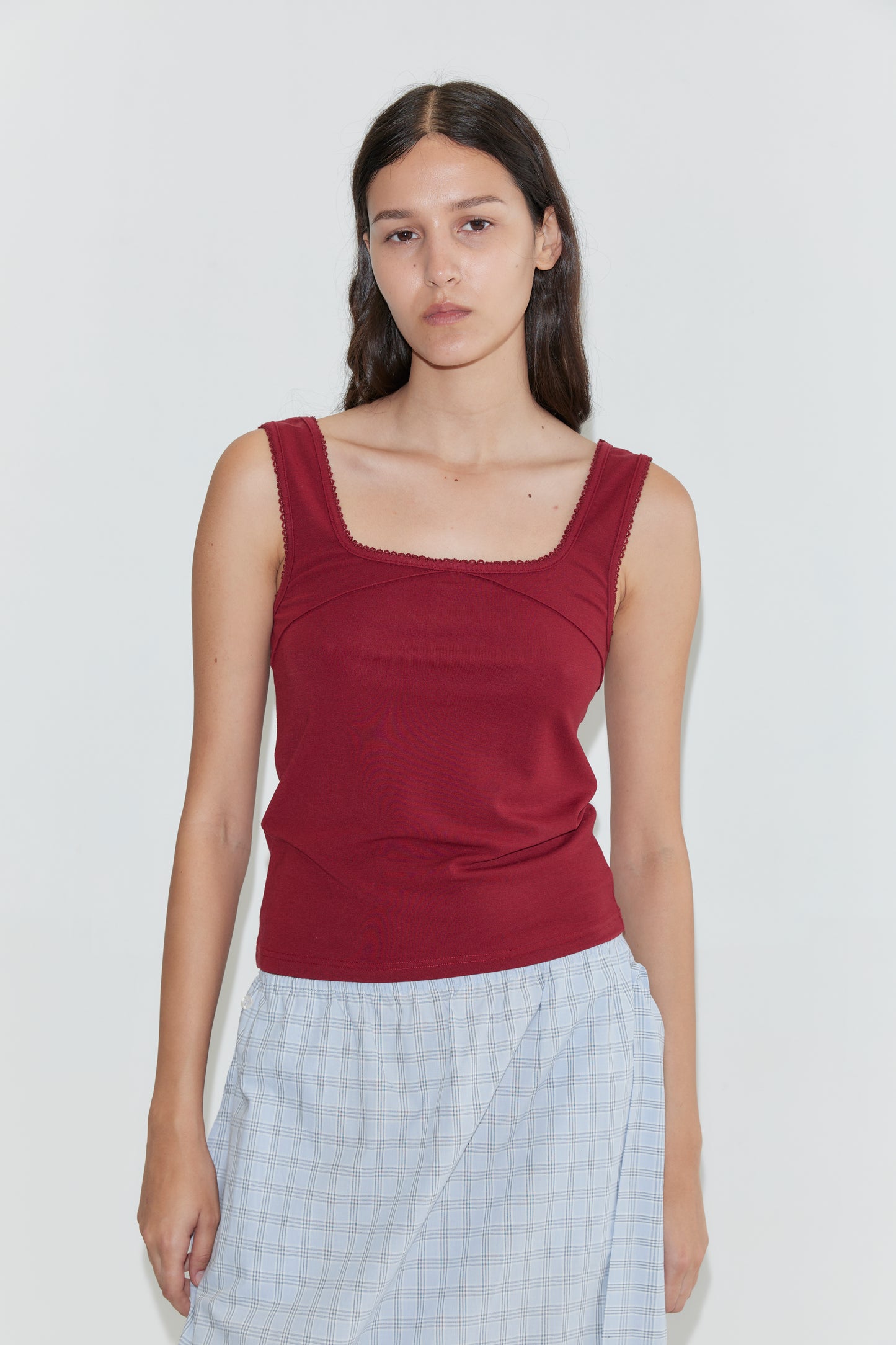 A woman with long dark hair wears the Piping Tank - Crimson, paired with a light blue plaid skirt. She stands against a plain white background, looking at the camera with a neutral expression.