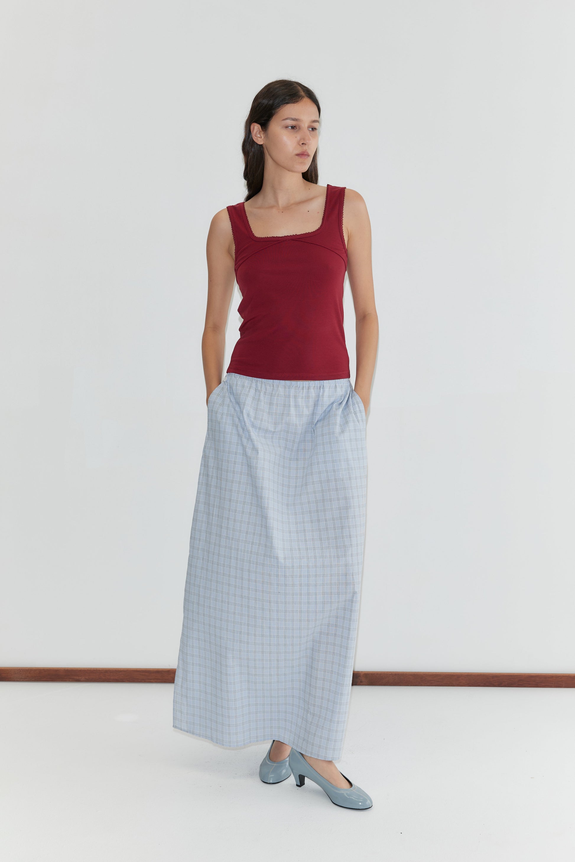 A woman with long dark hair stands against a white background, wearing a sleeveless maroon top and the Way Skirt - Powder Check by Deiji Studios, a light blue full-length skirt with pockets. She gazes to the side.