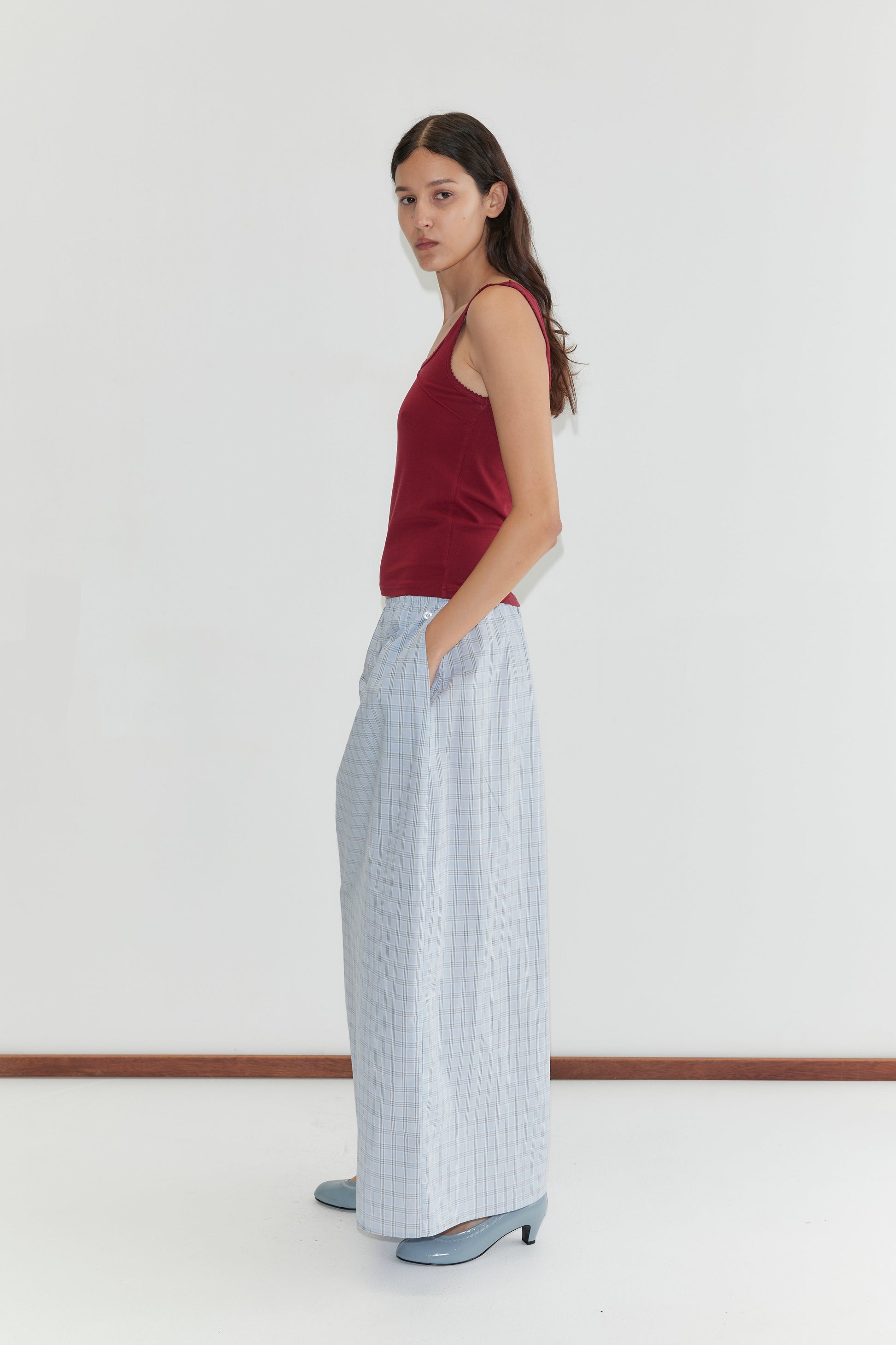 A woman stands sideways against a plain white background, wearing the Way Skirt - Powder Check by Deiji Studios, a sleeveless red top, and blue heels. She has long dark hair and her hands are tucked in her skirt pockets.