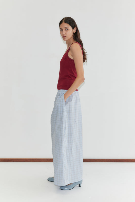 A woman stands sideways against a plain white background, wearing the Way Skirt - Powder Check by Deiji Studios, a sleeveless red top, and blue heels. She has long dark hair and her hands are tucked in her skirt pockets.