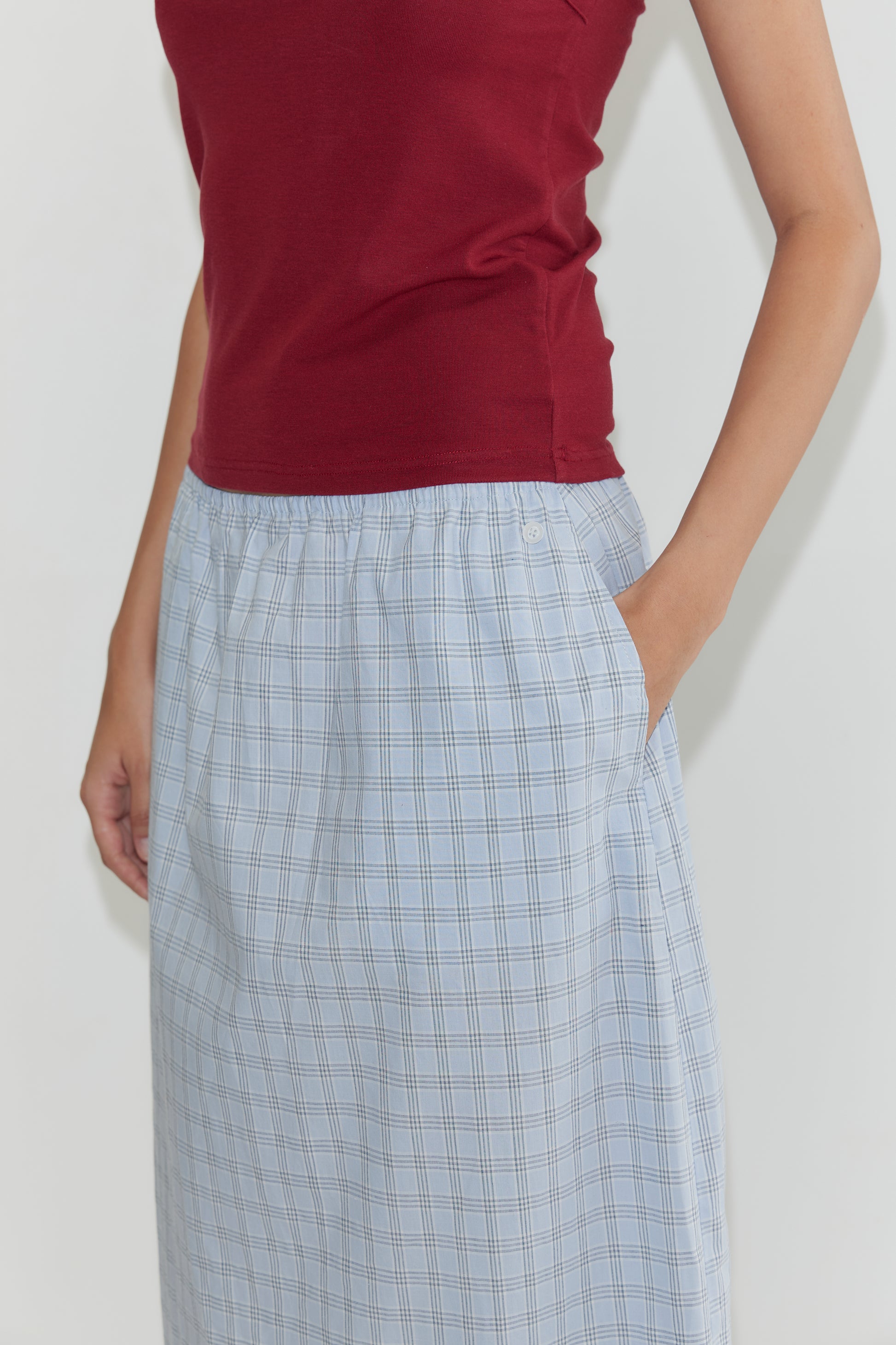 Someone wears a sleeveless burgundy top with the Way Skirt - Powder Check, an organic cotton light blue plaid skirt with pockets. Only the torso and upper legs are visible against a plain white background.