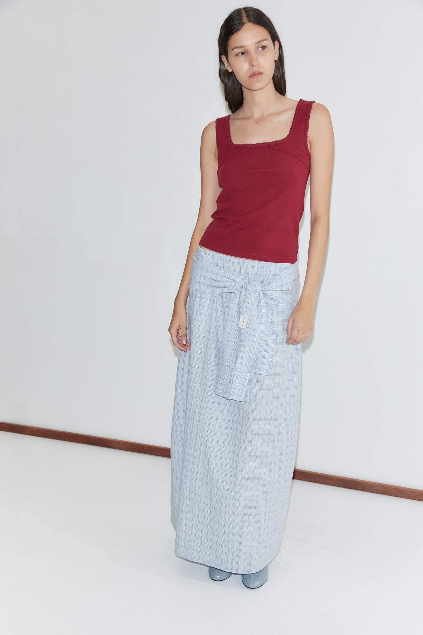 A woman with long dark hair and a neutral expression stands against a white background wearing the Way Skirt - Powder Check, a full-length light blue checkered skirt with a tied front detail from Deiji Studios and a sleeveless red top.