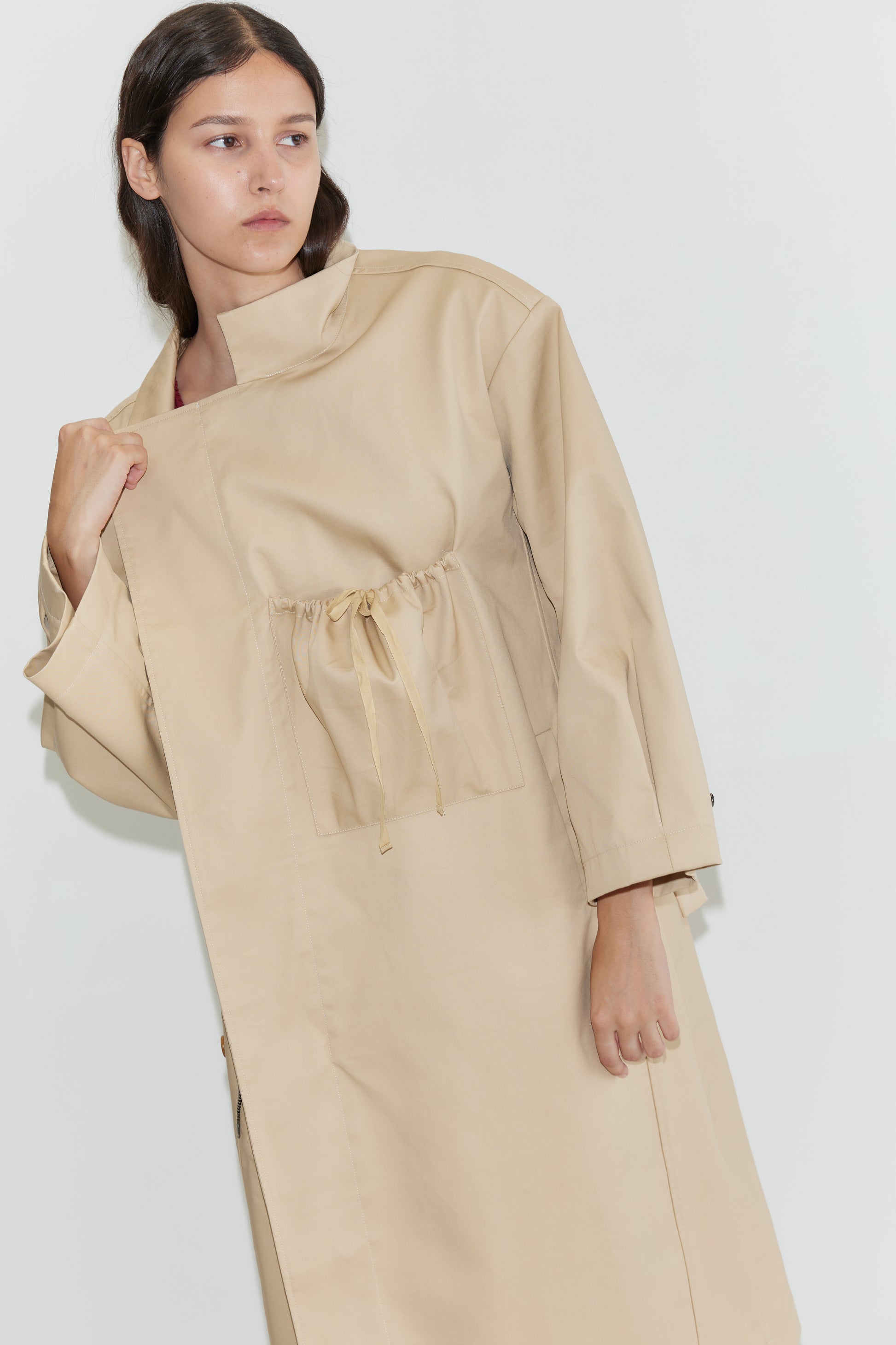 A woman with long brown hair stands against a plain white background wearing the Longline Jacket - Beige from Deiji Studios, featuring a loose fit, high collar, and drawstring waist.