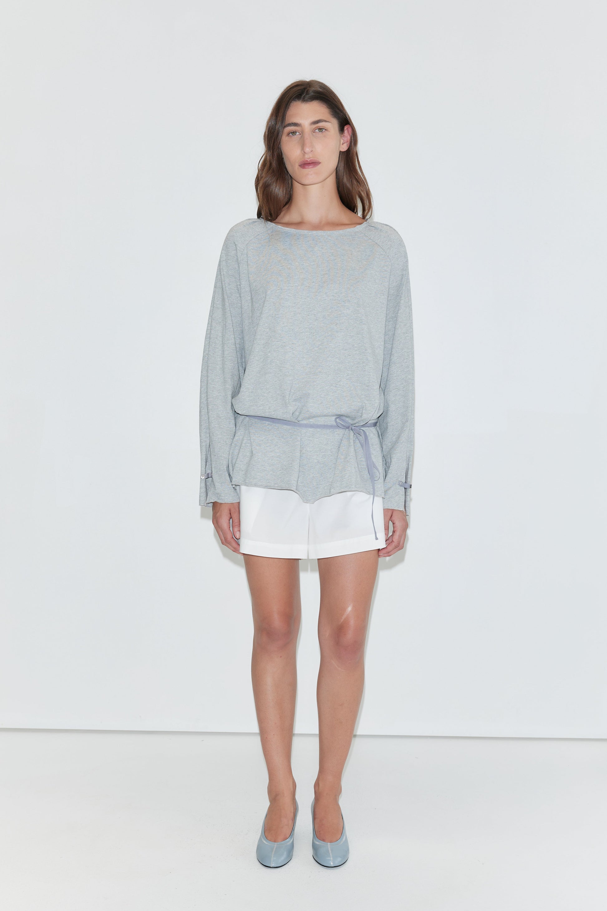 A woman with long dark hair stands against a plain white background wearing the Wided Longsleeve - Grey Marle by Deiji Studios, a grey marle jersey top with a wide neckline and drawstring hem, with white shorts and pale blue heels.