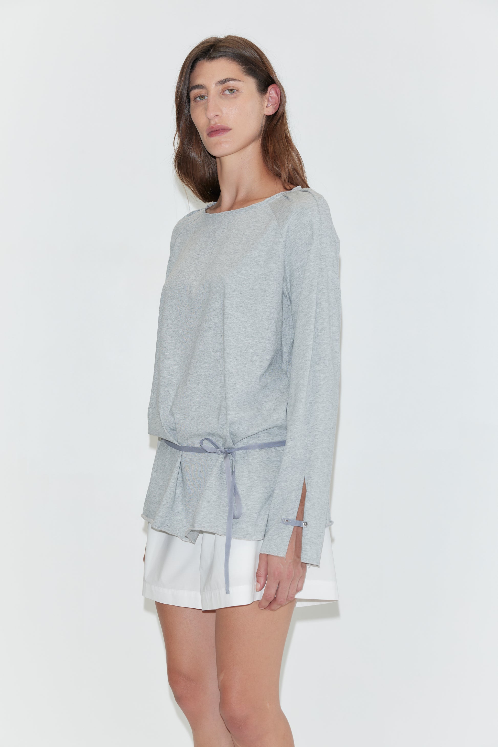 A woman with long dark hair stands in profile against a plain white background wearing the Wided Longsleeve - Grey Marle by Deiji Studios, a grey marle jersey top with a wide neckline and drawstring hem, with white shorts.