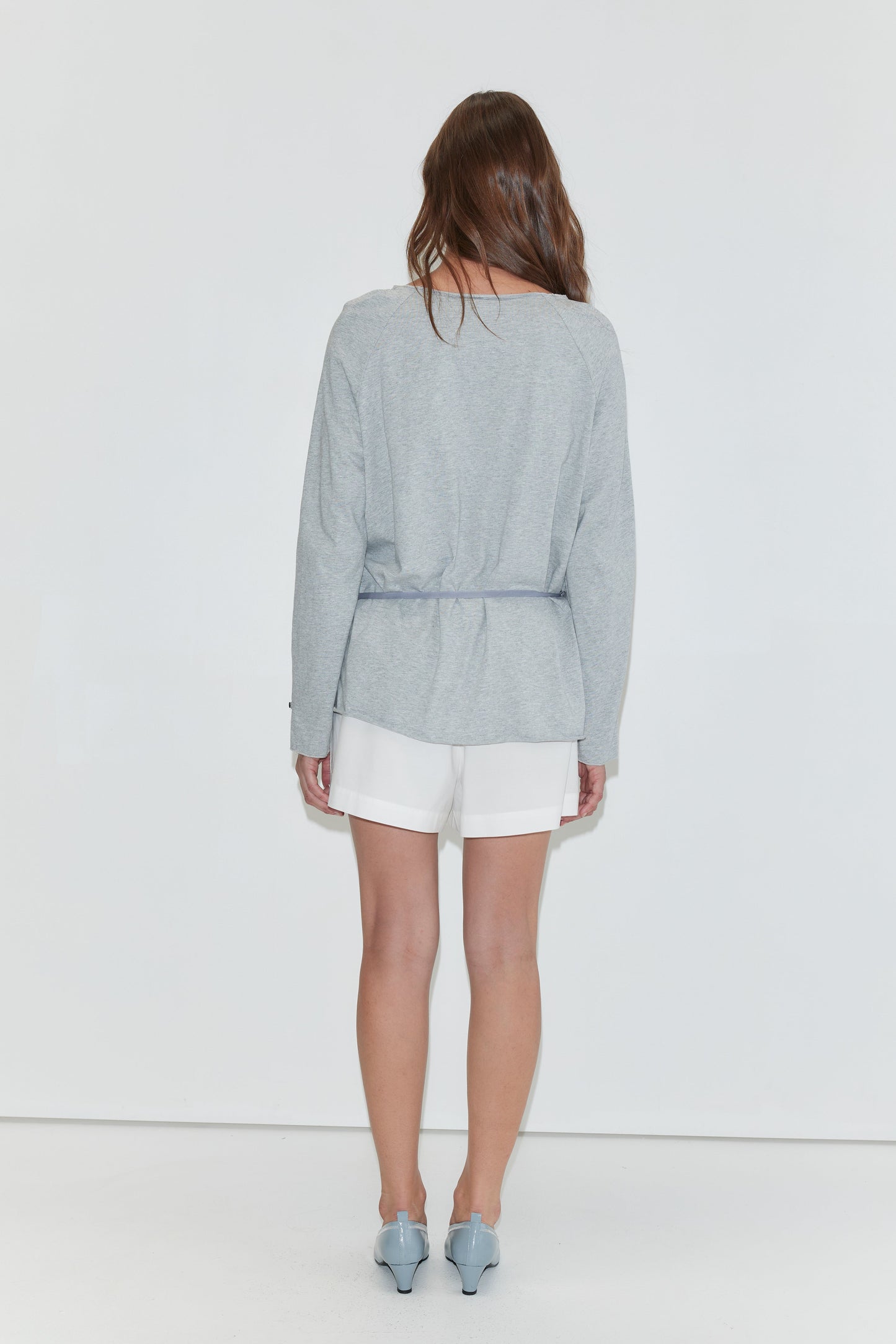 A woman with long dark hair stands with her back to the camera against a plain white background wearing the Wided Longsleeve - Grey Marle by Deiji Studios, a grey marle jersey top with a drawstring hem, with white shorts and pale blue heels.