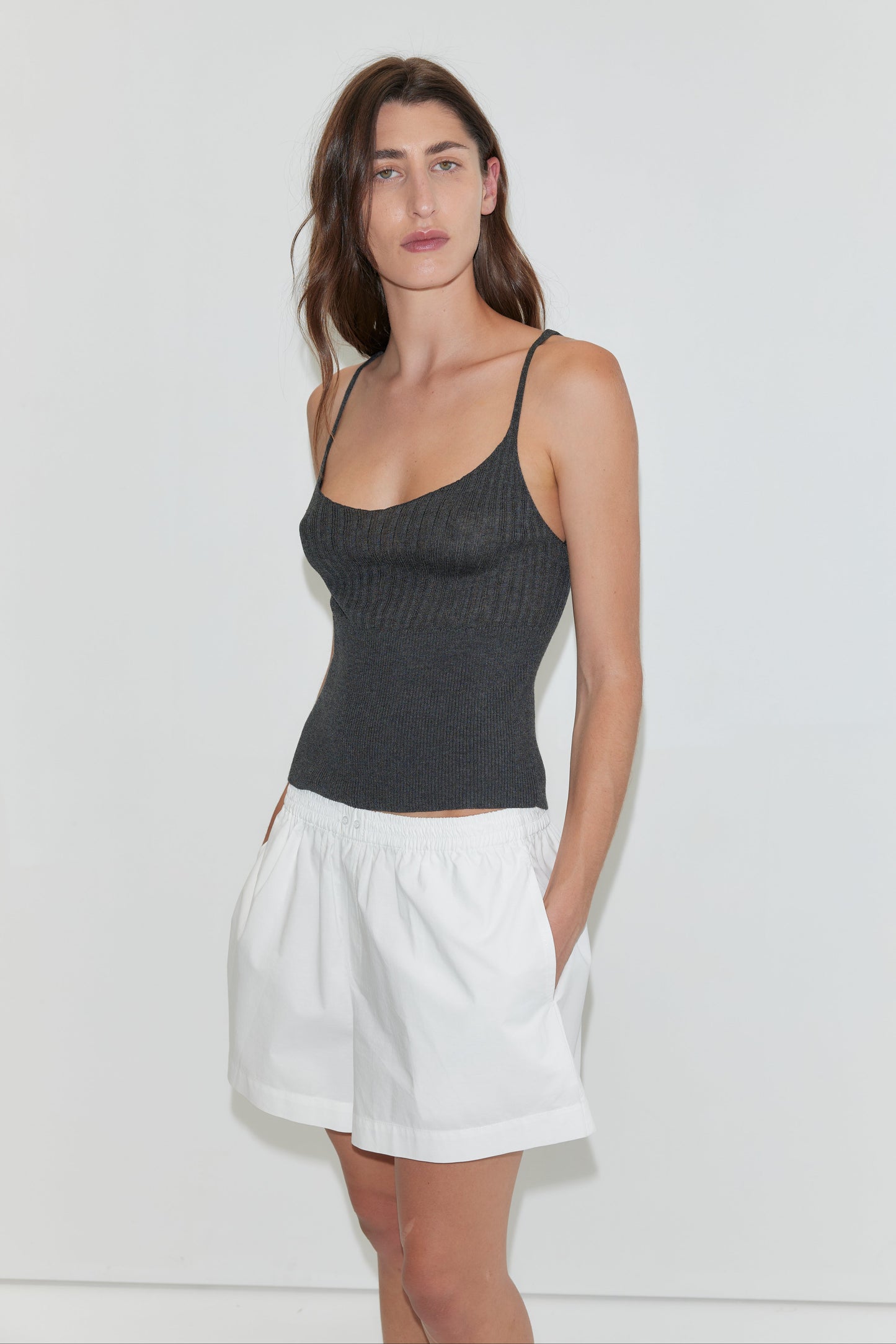 A woman with long brown hair poses against a white background, wearing a fitted black tank top and Deiji Studios Organic Cotton Boxer - White, her hands casually tucked in her pockets.
