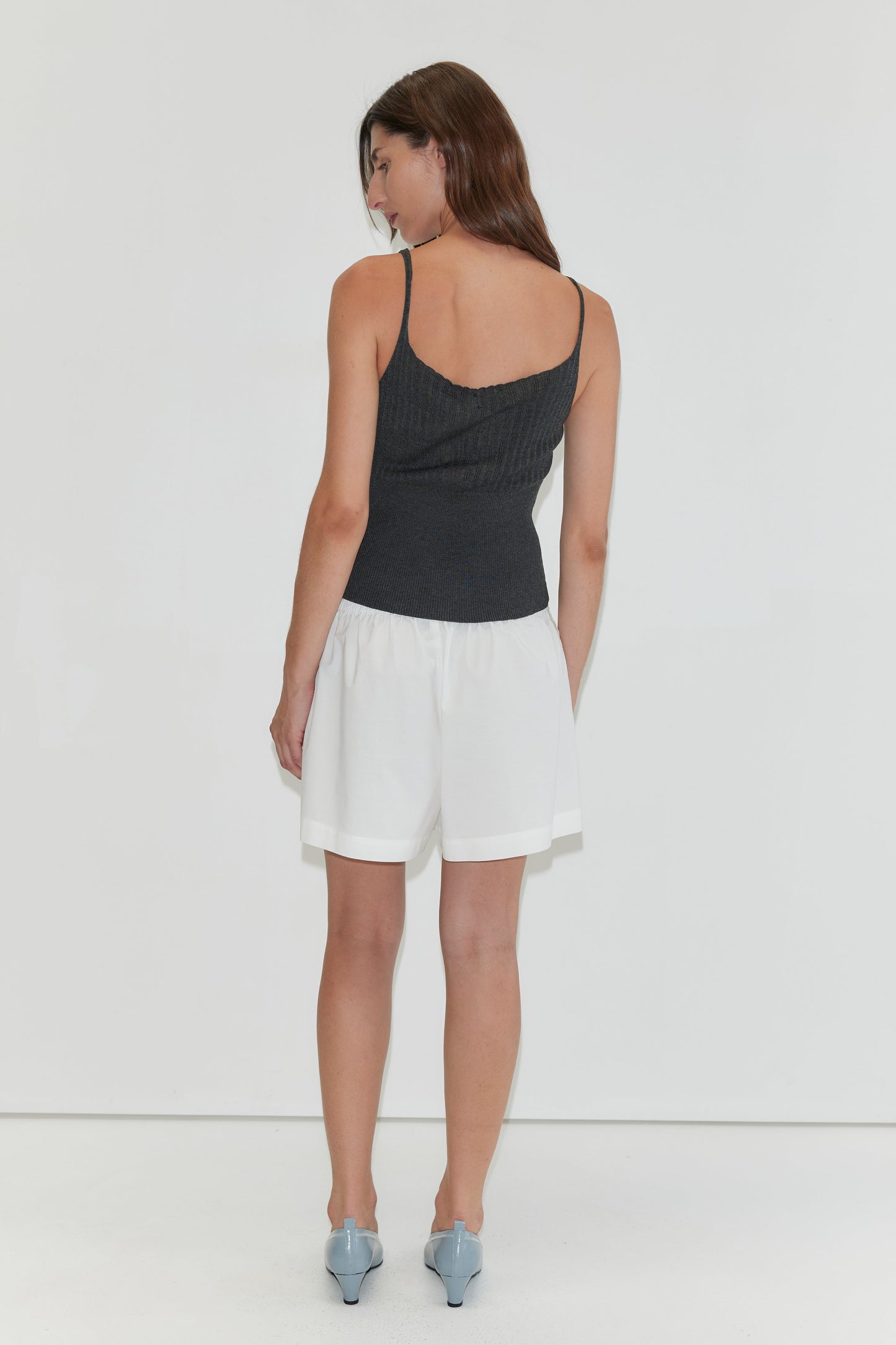 A woman faces away, wearing a black spaghetti strap top, Deiji Studios' Organic Cotton Boxer in white, and light blue pointed shoes, set against a plain white background.