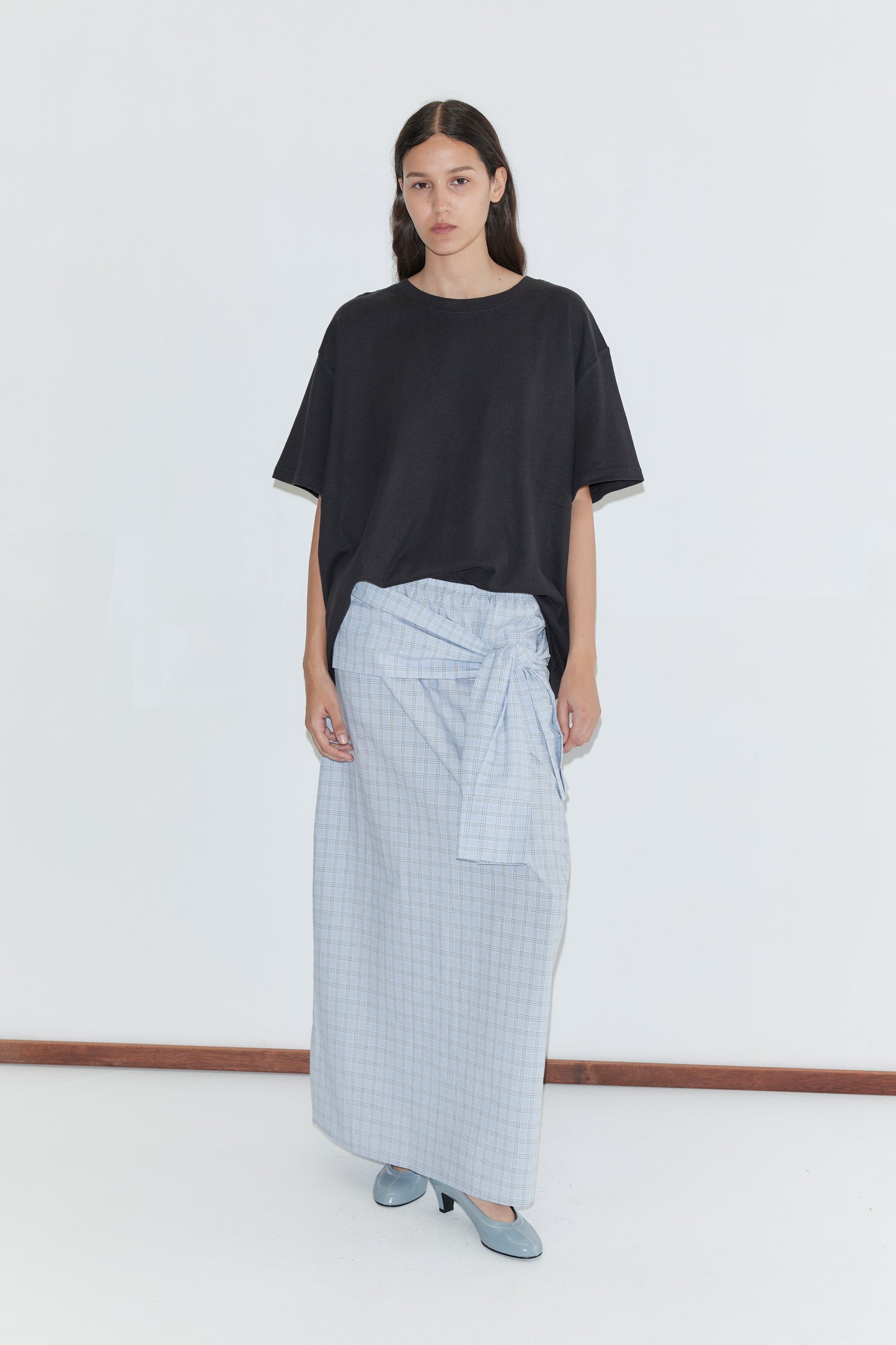 A woman stands against a plain white background, wearing a loose black T-shirt, blue heeled shoes, and the Shirt Cuff Belt - Powder Check—a light blue plaid wrap skirt with a cotton poplin belt. She has long dark hair and a neutral expression. Inspired by Deiji Studios style.