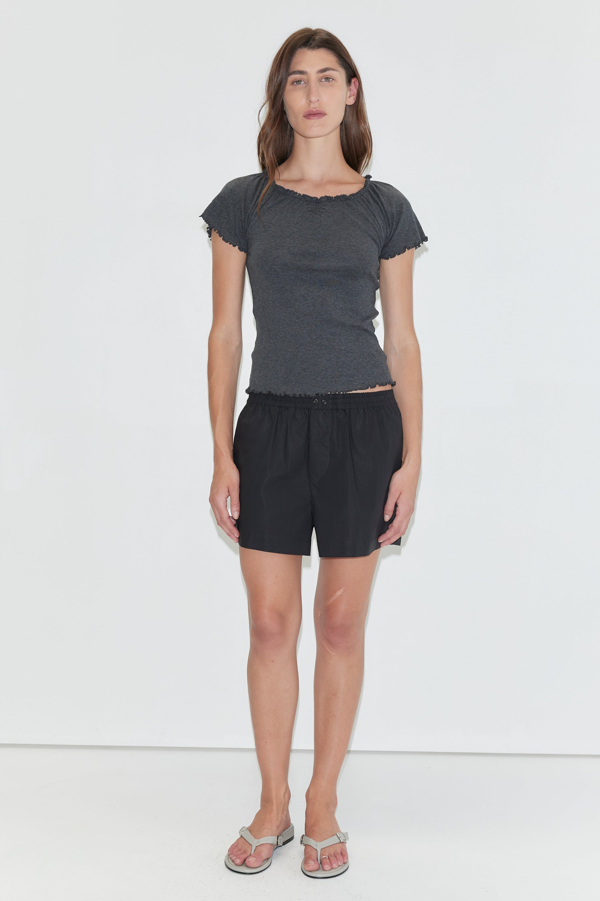 A person with long brown hair stands against a white background wearing a fitted dark gray short-sleeve top, light flip-flops, and Organic Cotton Boxer - Black—a loose, mid-rise boxer short with an elastic waist. They have a neutral expression and relaxed arms.