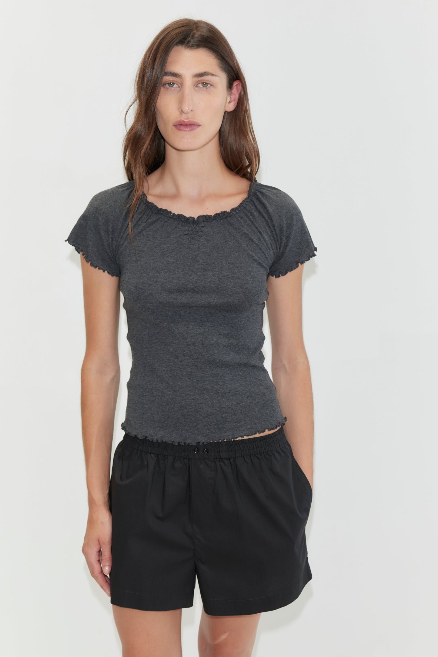 A woman with long brown hair stands against a plain white background, wearing loose black Organic Cotton Boxer shorts and a dark grey fitted short-sleeve top, with one hand in her pocket.