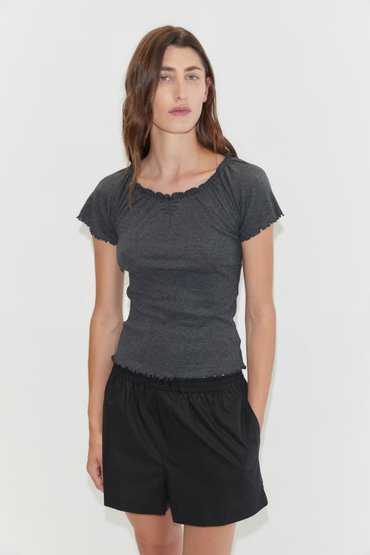 A woman with wavy brown hair wears a fitted dark gray top and loose Organic Cotton Boxer - Black shorts, standing against a plain white background with a neutral expression.