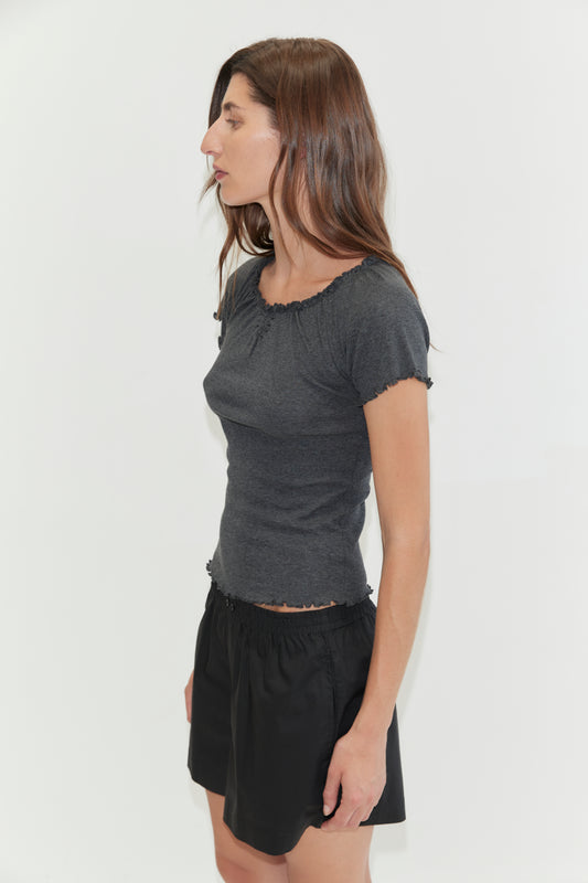 A woman stands in profile against a white background, wearing a fitted dark gray short-sleeve top and loose black Organic Cotton Boxer shorts. Her long brown hair is down, and her arms are relaxed at her sides.