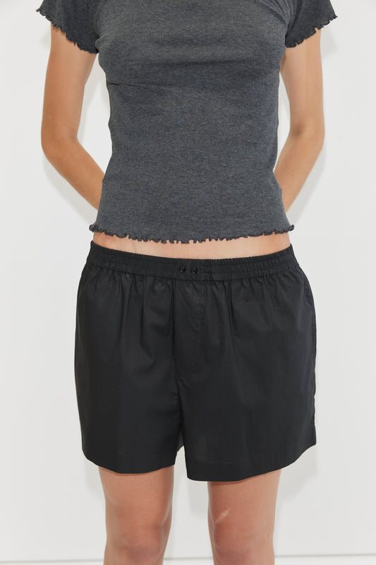Someone stands against a plain white background, arms behind their back, wearing loose black Organic Cotton Boxer shorts by Deiji Studios and a fitted, short-sleeved dark gray top.