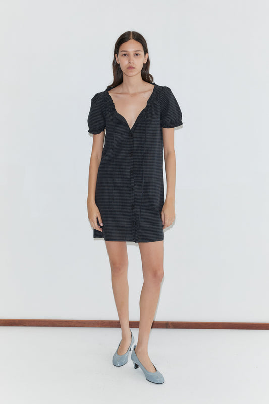A woman with long, straight hair stands against a plain white background, wearing the Buttoned Mini Dress - Mono Grid. The dress features short sleeves, a deep neckline, and is paired with light blue shoes.
