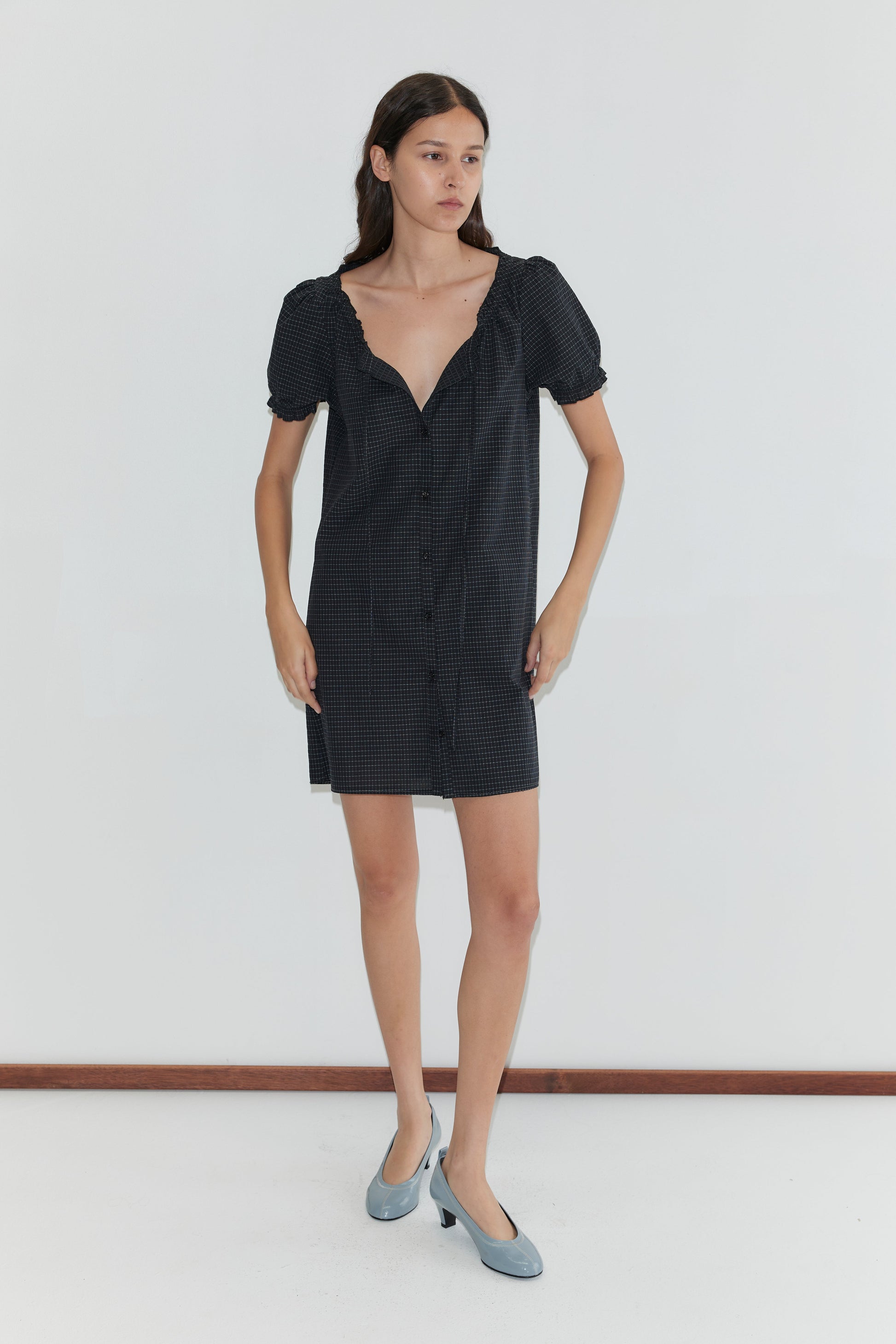 A woman with long brown hair stands against a plain white wall, wearing the Buttoned Mini Dress - Mono Grid and gray low-heeled shoes. She has a neutral expression and her hands rest by her sides.