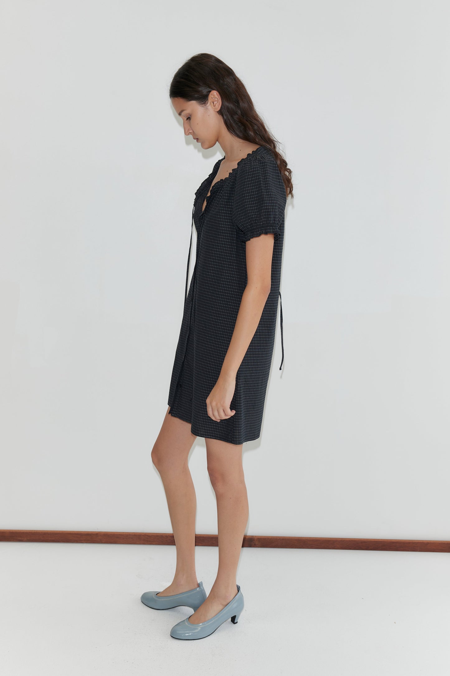 A woman with long brown hair wears the Buttoned Mini Dress - Mono Grid. The short dress has short sleeves and a dark grid pattern. She pairs it with light blue shoes and stands sideways, looking down, against a plain white background.