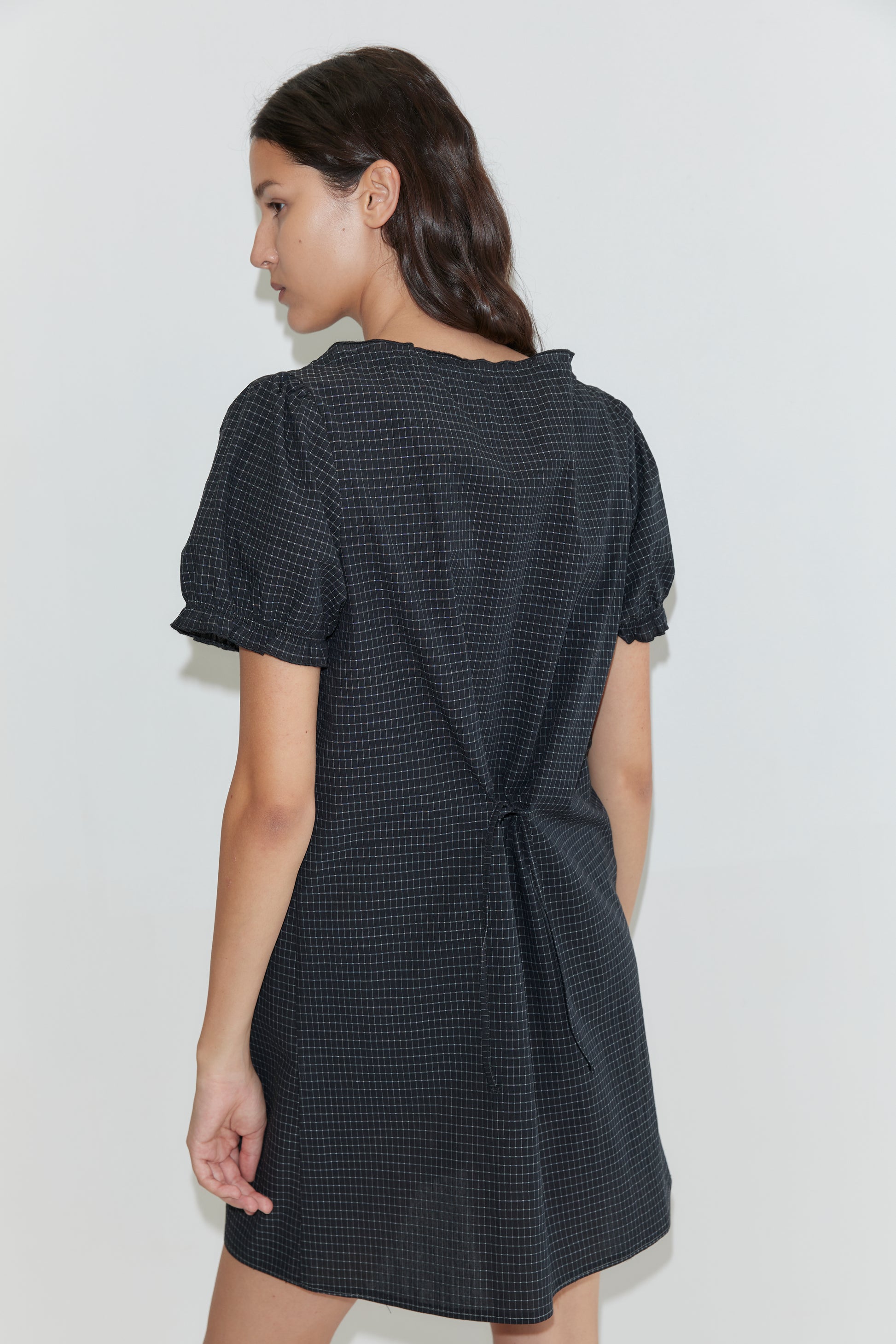 A woman with long brown hair stands facing away from the camera against a white background, wearing the Buttoned Mini Dress - Mono Grid, a short-sleeved shift dress in a dark checkered pattern with back tie detail.