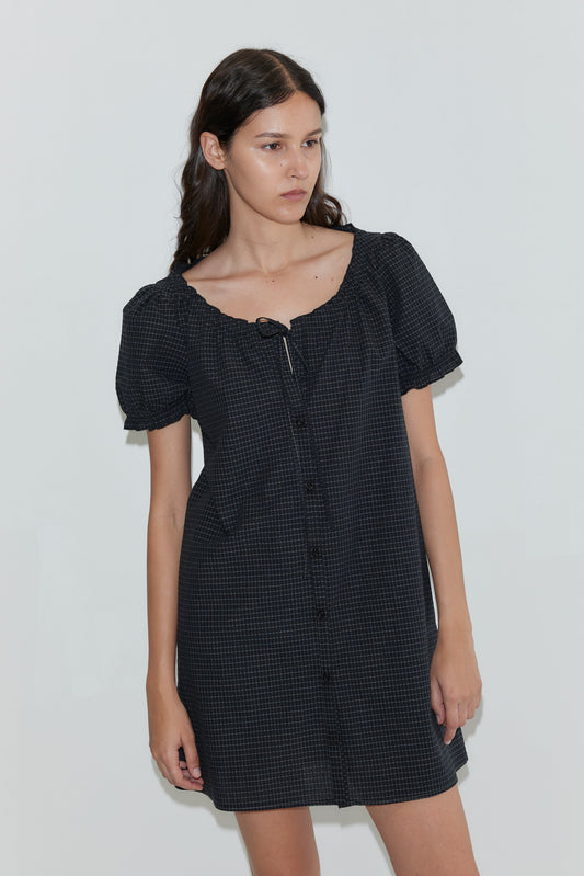 A woman with long dark hair wears the Buttoned Mini Dress - Mono Grid, featuring puffed short sleeves, standing against a plain light background.