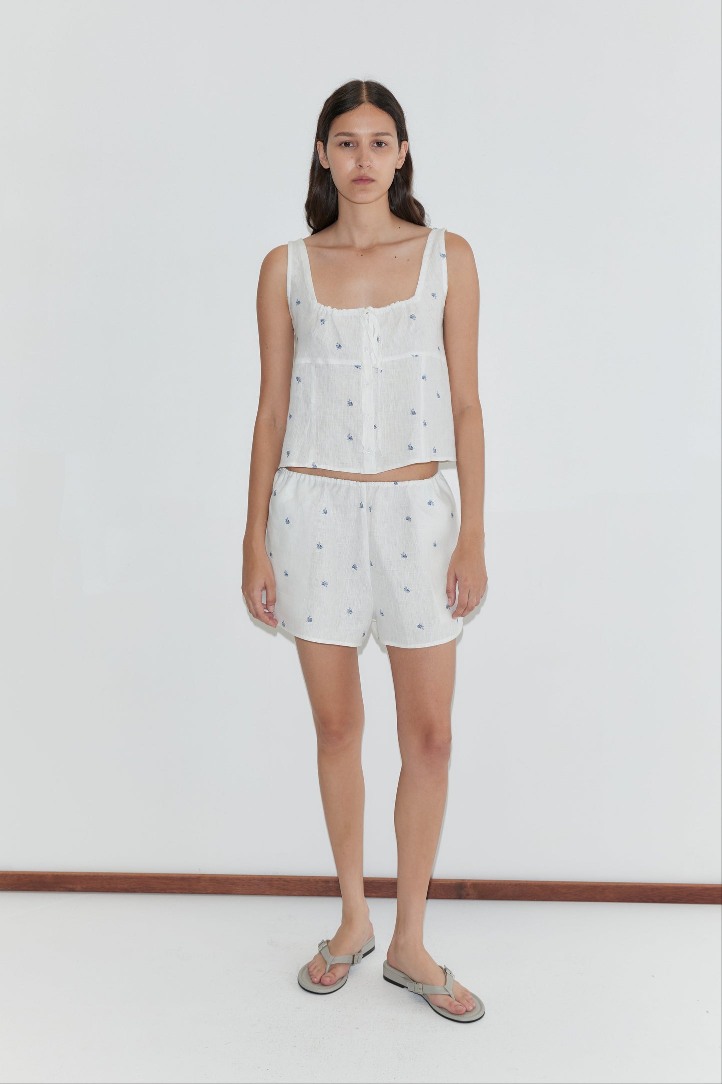 A woman with long brown hair and a neutral expression stands against a white background, wearing the Deiji Studios Relax Short Set - Floral Blue and gray flip-flops.