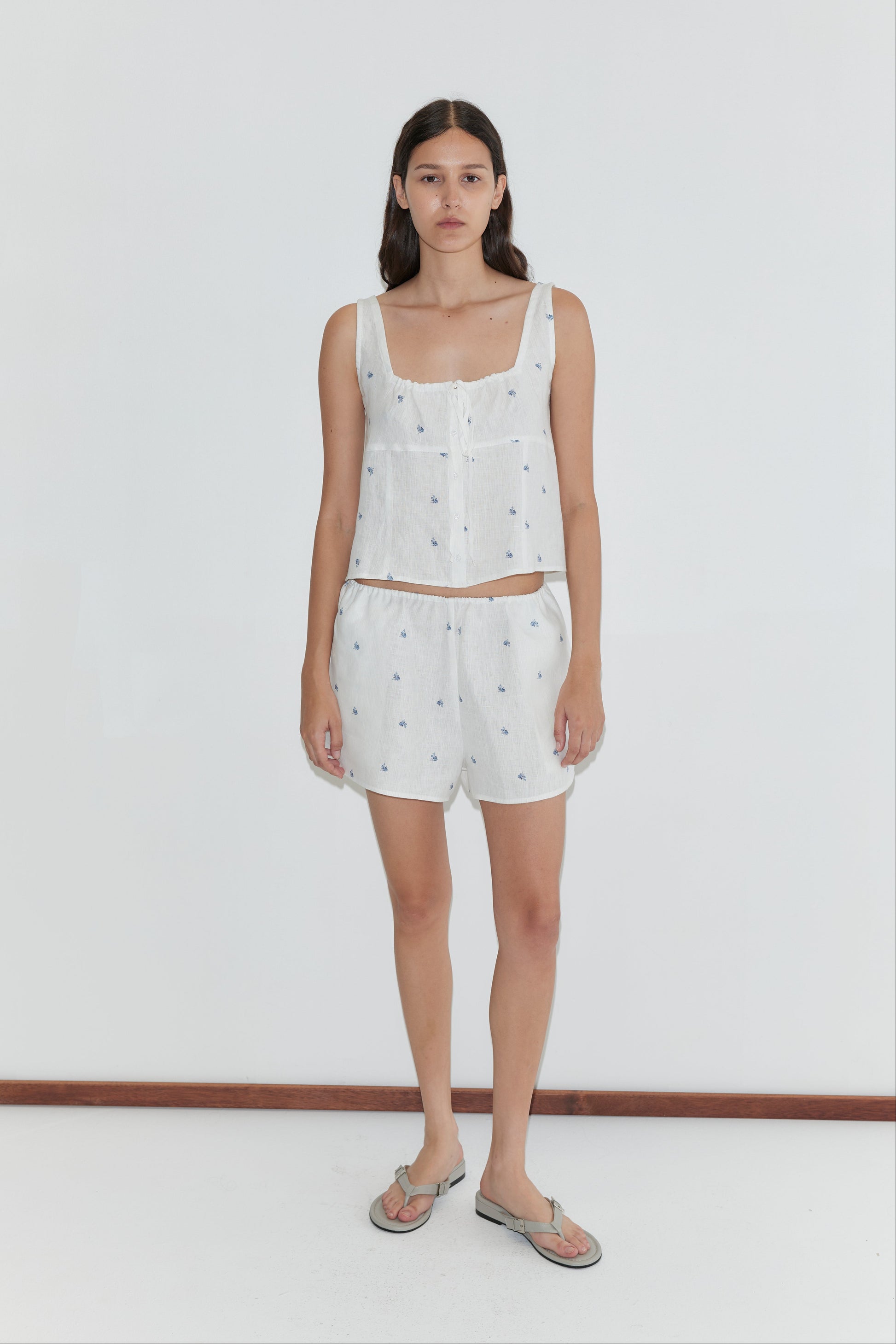 A woman with long brown hair and a neutral expression stands against a white background, wearing the Deiji Studios Relax Short Set - Floral Blue and gray flip-flops.