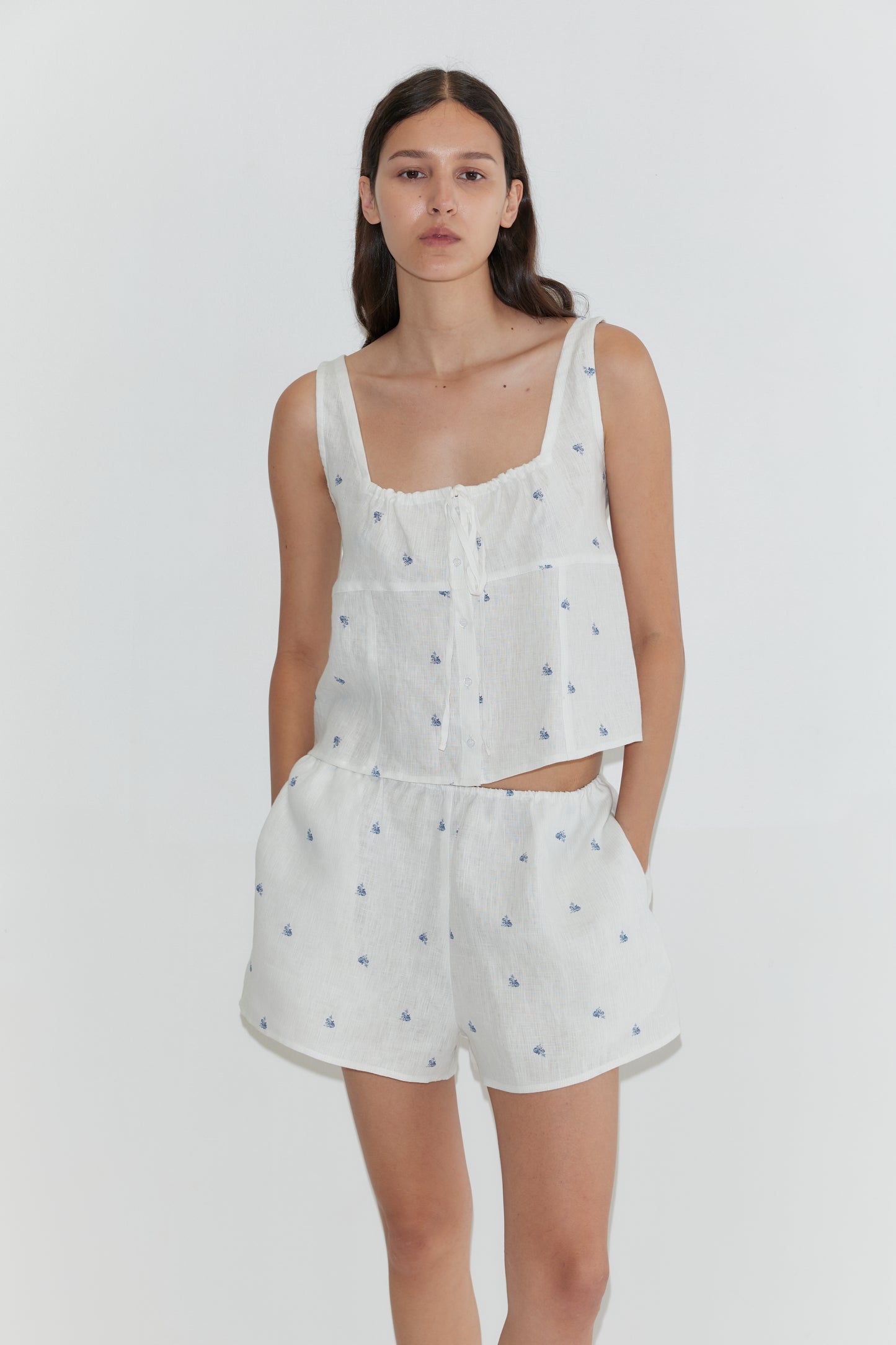 A woman wears the Relax Short Set - Floral Blue by Deiji Studios, featuring a sleeveless white linen top and matching shorts with blue floral prints, standing against a plain white background.
