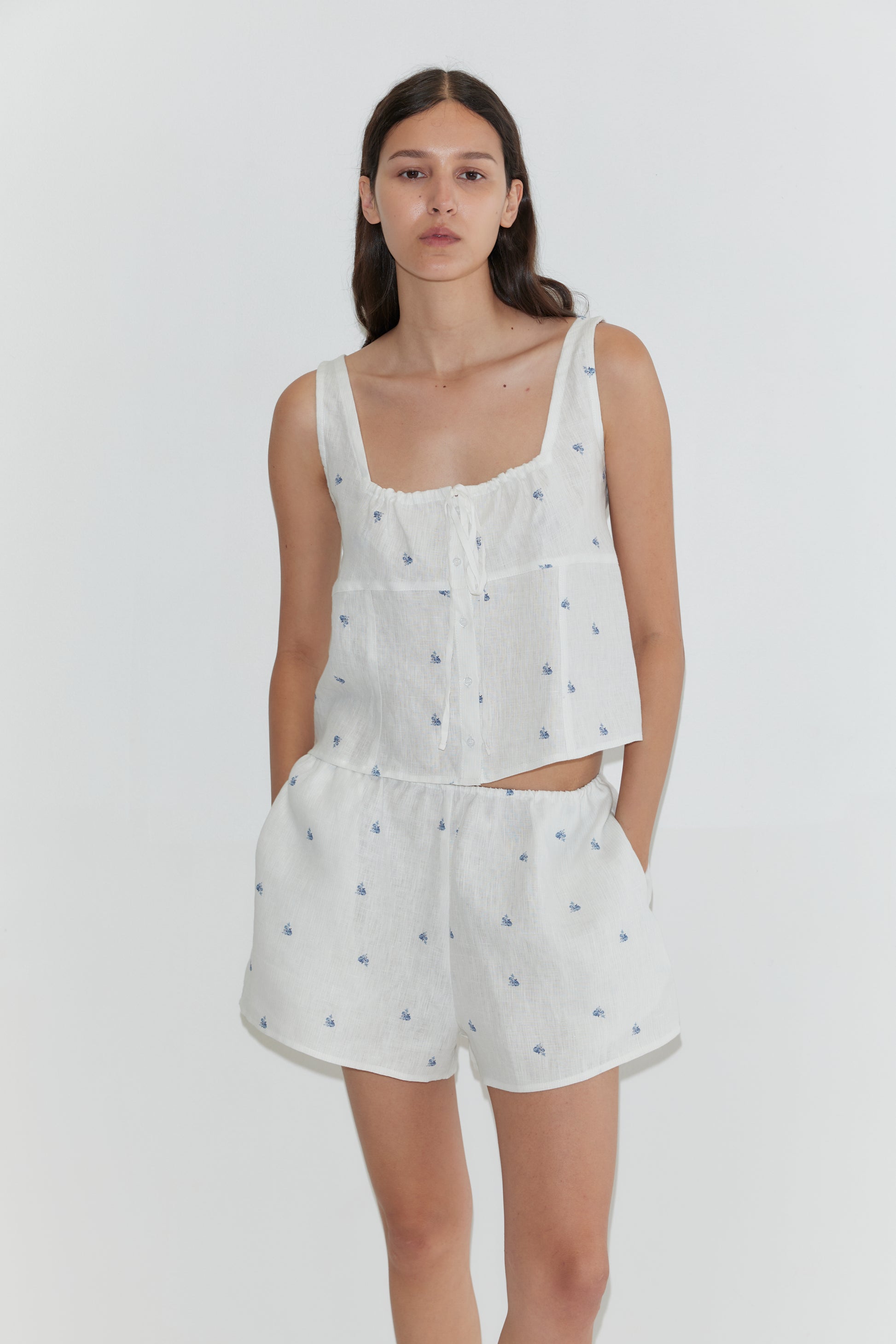 A woman wears the Relax Short Set - Floral Blue by Deiji Studios, featuring a sleeveless white linen top and matching shorts with blue floral prints, standing against a plain white background.