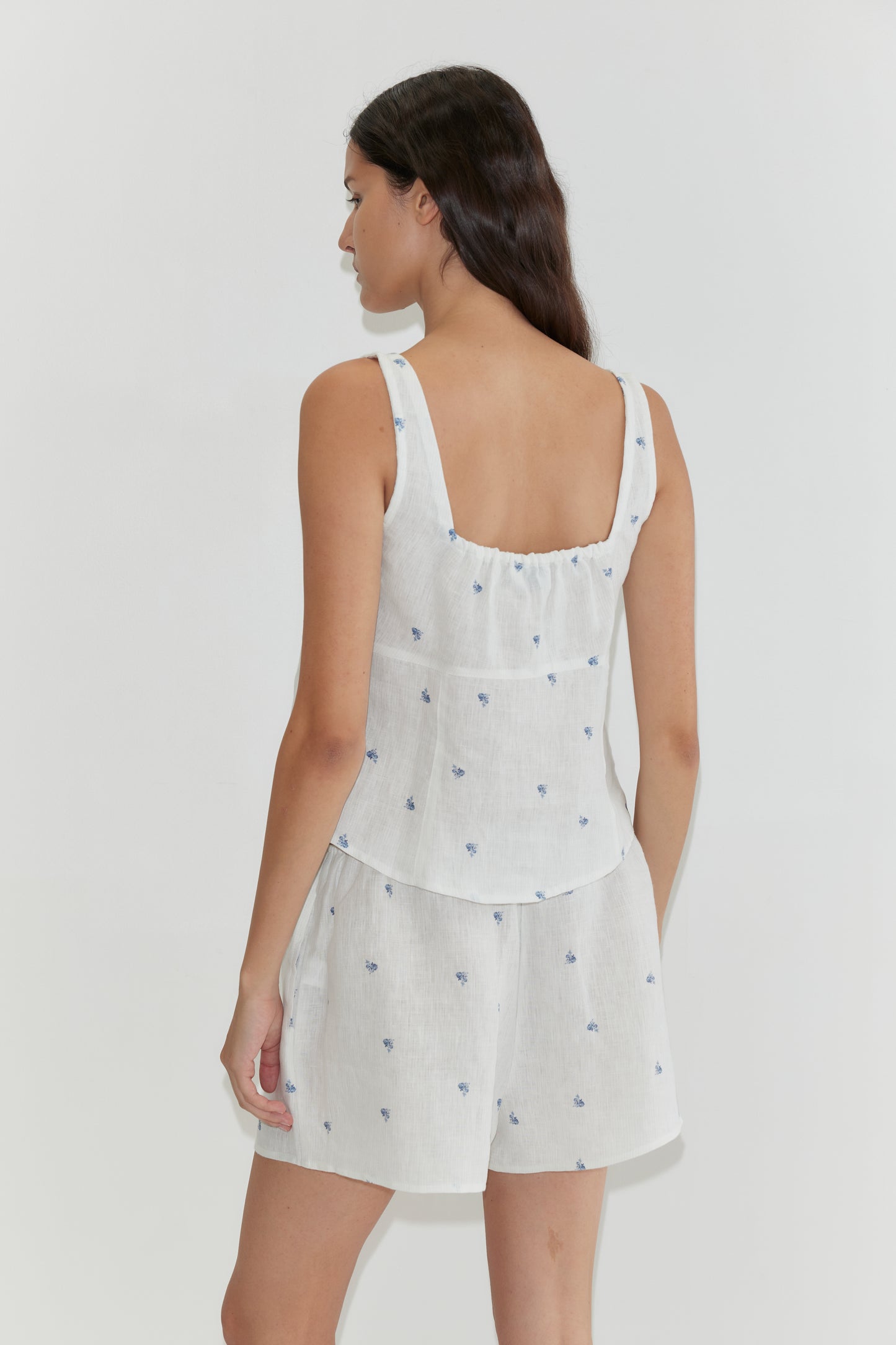 A woman with long dark hair faces away, wearing the Relax Short Set - Floral Blue by Deiji Studios, featuring a subtle blue floral pattern on white linen, set against a plain light background.