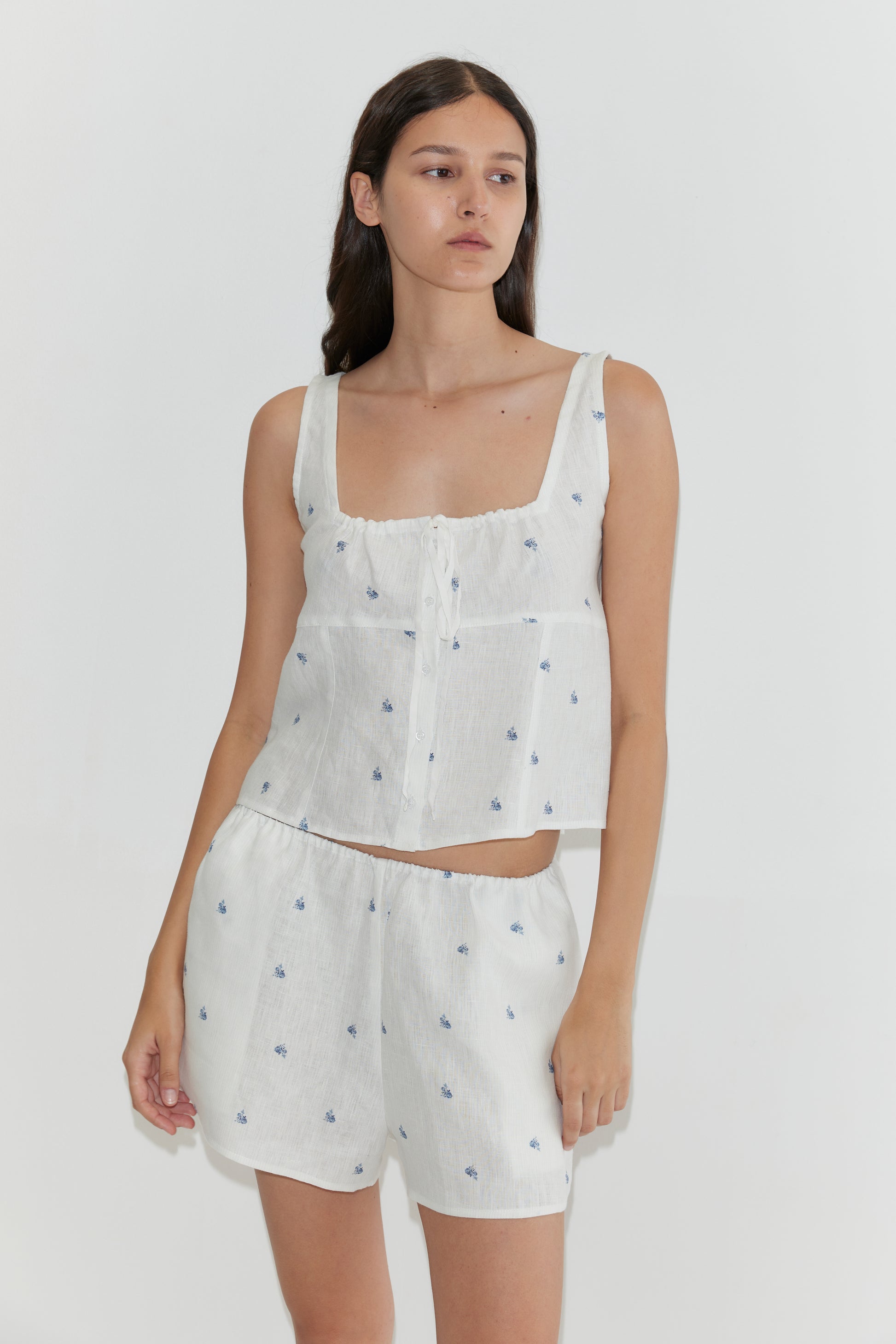 A woman wears the Deiji Studios Relax Short Set - Floral Blue, featuring a white linen tank and shorts with small blue floral patterns, standing against a plain white background.