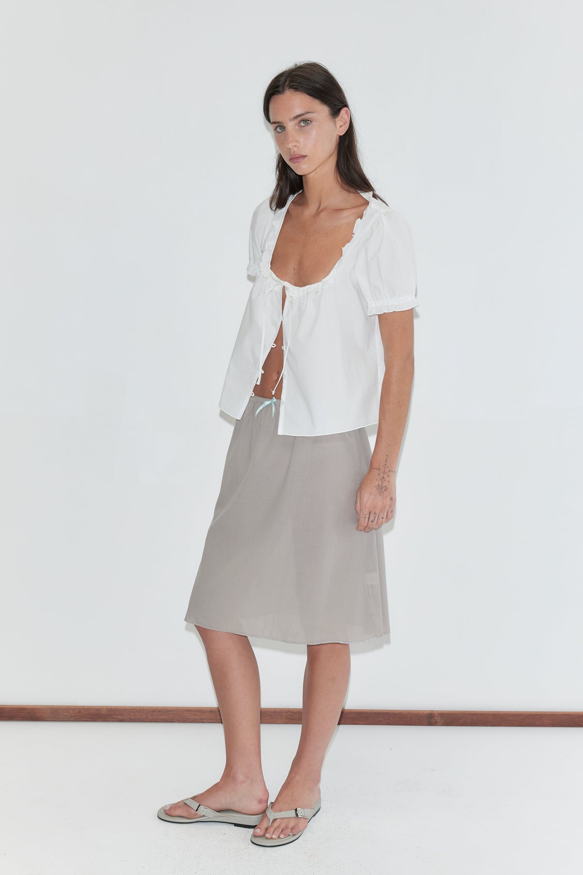A woman with long dark hair stands against a plain white background wearing an open white frill top and the Basic Skirt - Grey by Deiji Studios, a grey cotton voile midi skirt, with grey flip-flops.