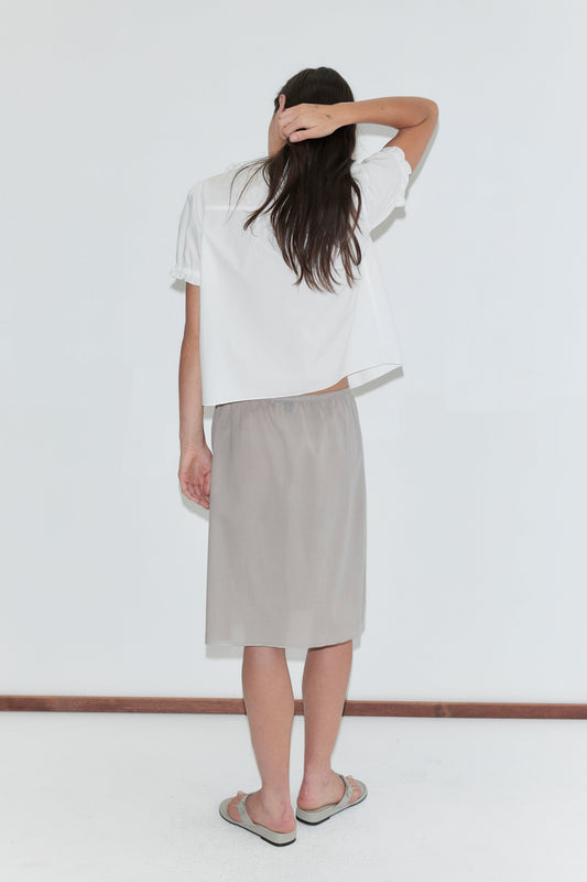 A woman with long dark hair stands with her back to the camera against a plain white background, wearing a white frill top and the Basic Skirt - Grey by Deiji Studios, a grey cotton voile midi skirt, with grey flip-flops.