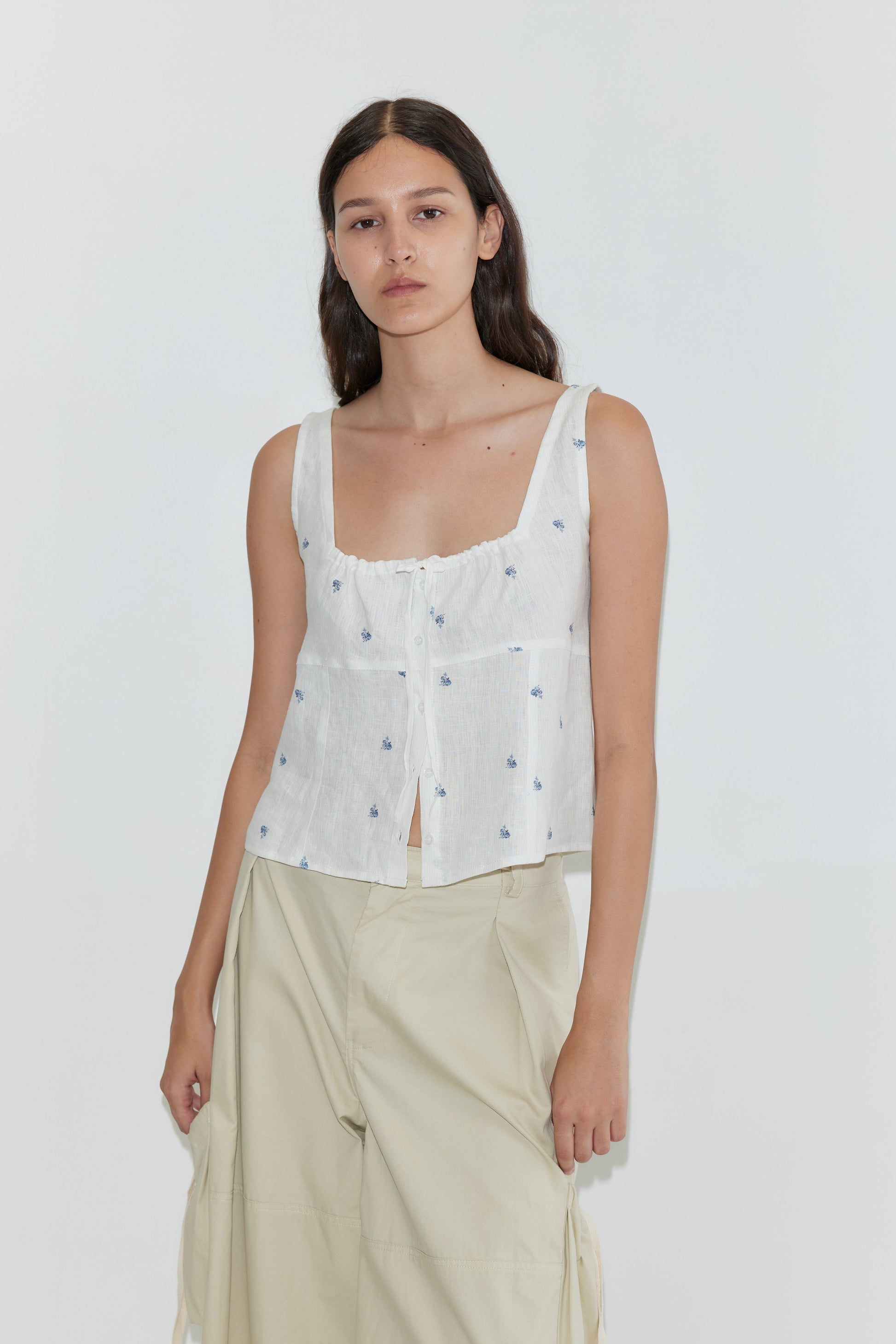 A woman with long dark hair and a neutral expression models the Relax Tie Top - Floral Blue by Deiji Studios, featuring a white sleeveless linen design with blue florals, paired with light beige wide-leg pants against a plain background.