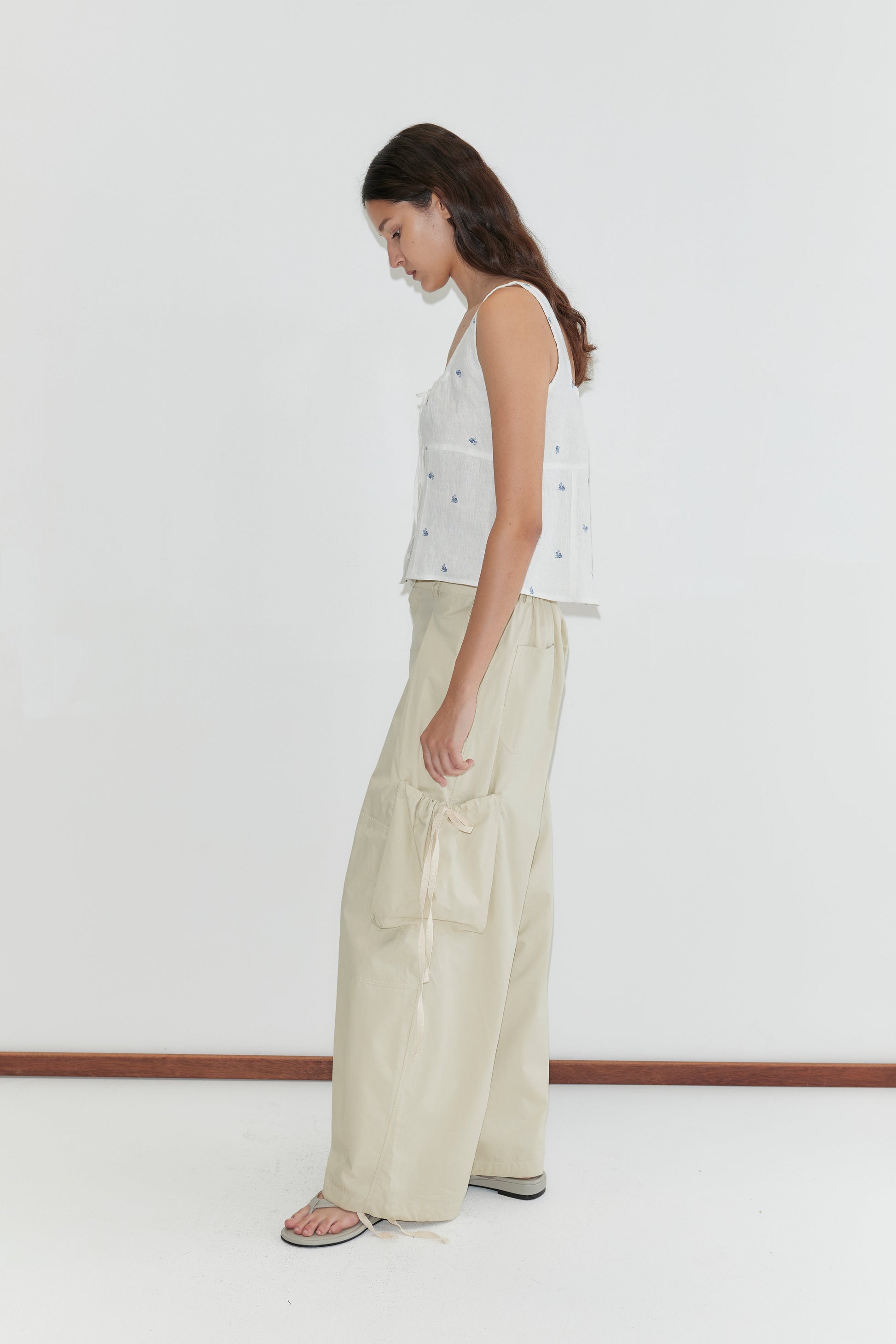 A woman with long brown hair stands sideways, barefoot on a white floor against a plain wall, wearing the Deiji Studios Relax Tie Top in Floral Blue paired with loose, light beige pants with large pockets.