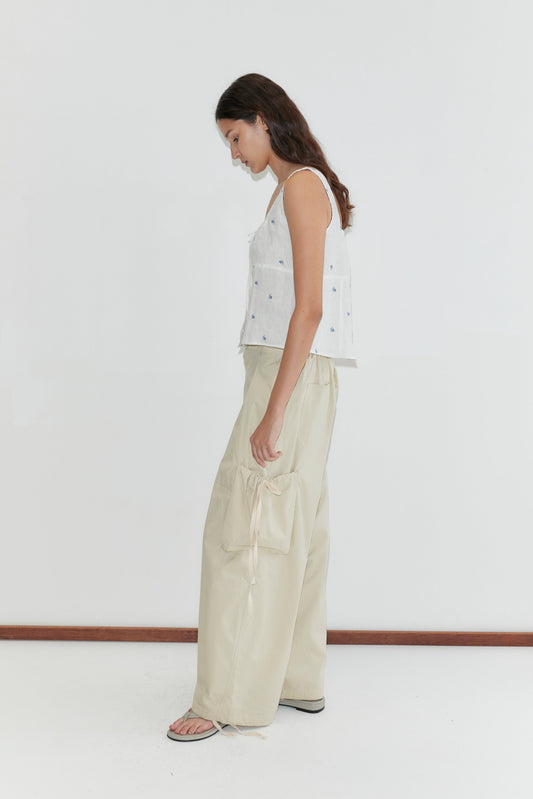 A woman with long brown hair stands sideways, barefoot on a white floor against a plain wall, wearing the Deiji Studios Relax Tie Top in Floral Blue paired with loose, light beige pants with large pockets.