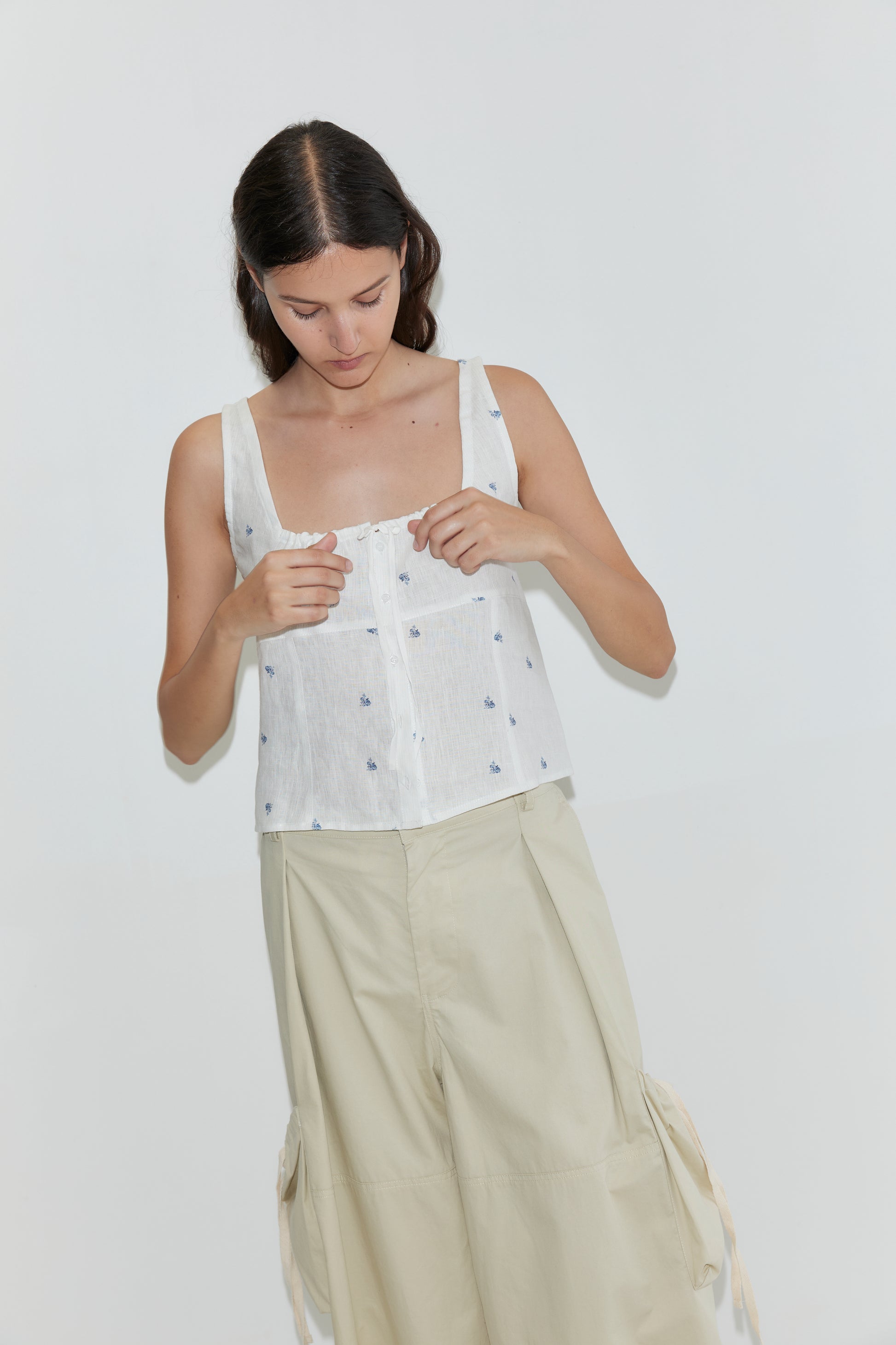 A woman with long dark hair looks down as she fastens buttons on the Relax Tie Top - Floral Blue, a sleeveless white linen tank with subtle blue floral patterns. She pairs it with wide-leg beige pants; the background is plain and light-colored.