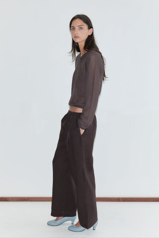 A woman with long dark hair stands in profile against a plain white background with a hand in her pocket, wearing the Linen Ease Trouser - Brown Black Stripe by Deiji Studios, dark brown fine-striped wide-leg linen trousers, with a matching brown striped knit top and pale blue heels.