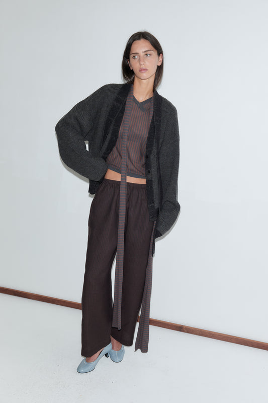 A woman with long dark hair stands against a plain white background wearing the Linen Ease Trouser - Brown Black Stripe by Deiji Studios, dark brown fine-striped wide-leg linen trousers, with a brown striped knit top, a charcoal cardigan and pale blue heels.