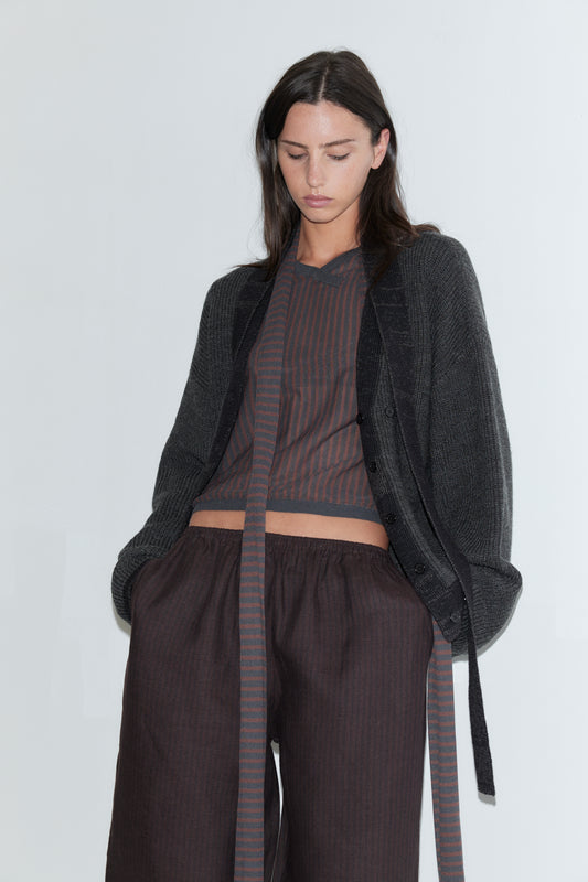 A cropped view of a woman wearing the Linen Ease Trouser - Brown Black Stripe by Deiji Studios, dark brown fine-striped wide-leg linen trousers, with a brown striped knit top and an open charcoal cardigan against a plain white background.