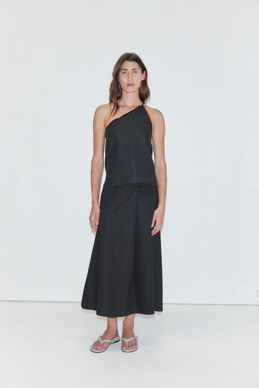 A person stands against a plain white background wearing a black one-shoulder sleeveless top, the Wrap Skirt - Black, and gray flip-flops. They have long brown hair and a neutral expression.