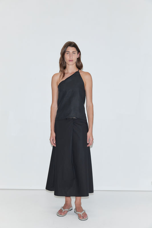 A person stands against a plain white background, wearing a black one-shoulder top and the Wrap Skirt - Black from Deiji Studios, paired with sandals.