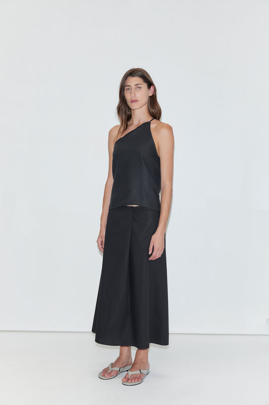 A woman stands against a plain white background wearing the Deiji Studios Half Halter - Black, paired with a black wide-leg skirt and silver flip-flops. She has long brown hair and a relaxed, neutral expression.