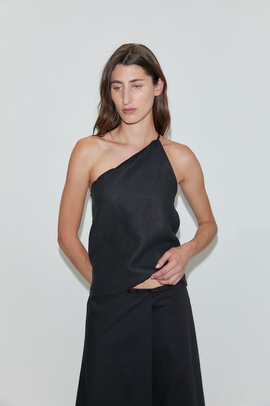 A woman with long brown hair wears the Wrap Skirt - Black by Deiji Studios, a sleeveless one-shoulder black top, and stands against a plain white background.