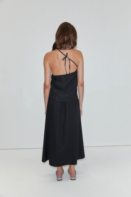 A woman with long brown hair stands facing away, wearing the Wrap Skirt - Black from Deiji Studios, a flowing halter-neck dress crafted in soft cotton poplin. She completes the look with gray heels against a plain white background.