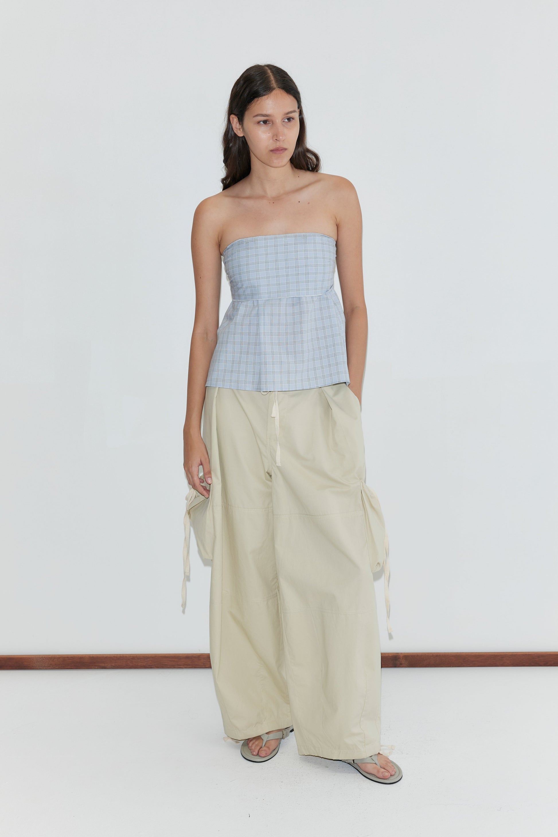 A woman with long dark hair stands against a white background wearing the Deiji Studios Flow Top in Powder Check, paired with loose beige wide-leg pants with side ties, sandals, and her hands in her pockets.