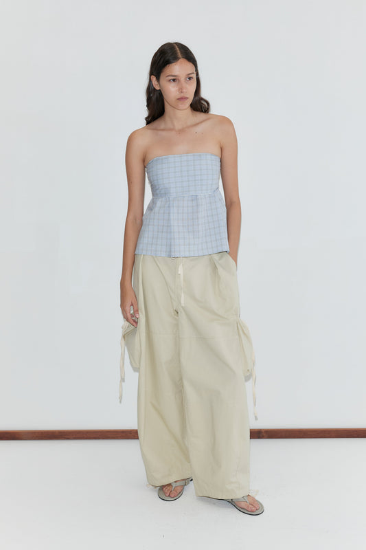 A woman with long dark hair stands against a white background wearing the Deiji Studios Flow Top in Powder Check, paired with loose beige wide-leg pants with side ties, sandals, and her hands in her pockets.