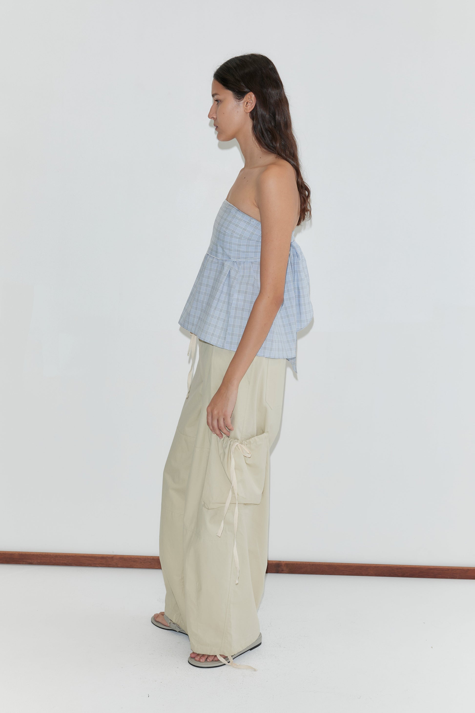 A woman stands in profile against a white background, wearing the Flow Top - Powder Check by Deiji Studios, a strapless light blue plaid cotton top, loose beige cargo pants with side pockets and drawstrings, and black sandals.