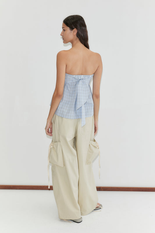 A woman stands against a white background, facing away. She wears the Deiji Studios Flow Top - Powder Check, featuring a strapless design with a back bow and removable scarf panel, paired with loose beige cargo pants with large pockets.