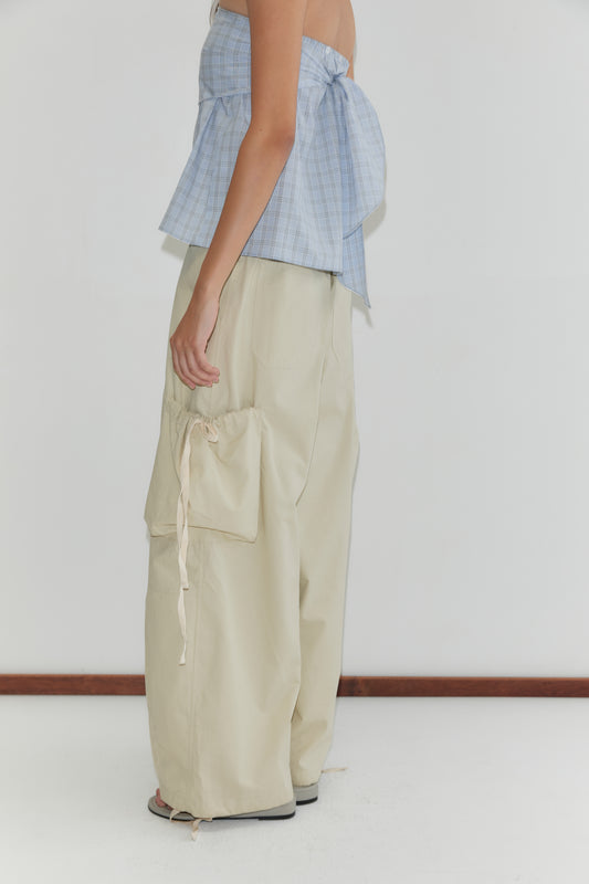 A person stands sideways against a white background, wearing a blue plaid strapless top with a large bow and high-waisted Deiji Studios Cargo Pant - Stone, featuring wide legs and an oversized pocket with ribbon tie.