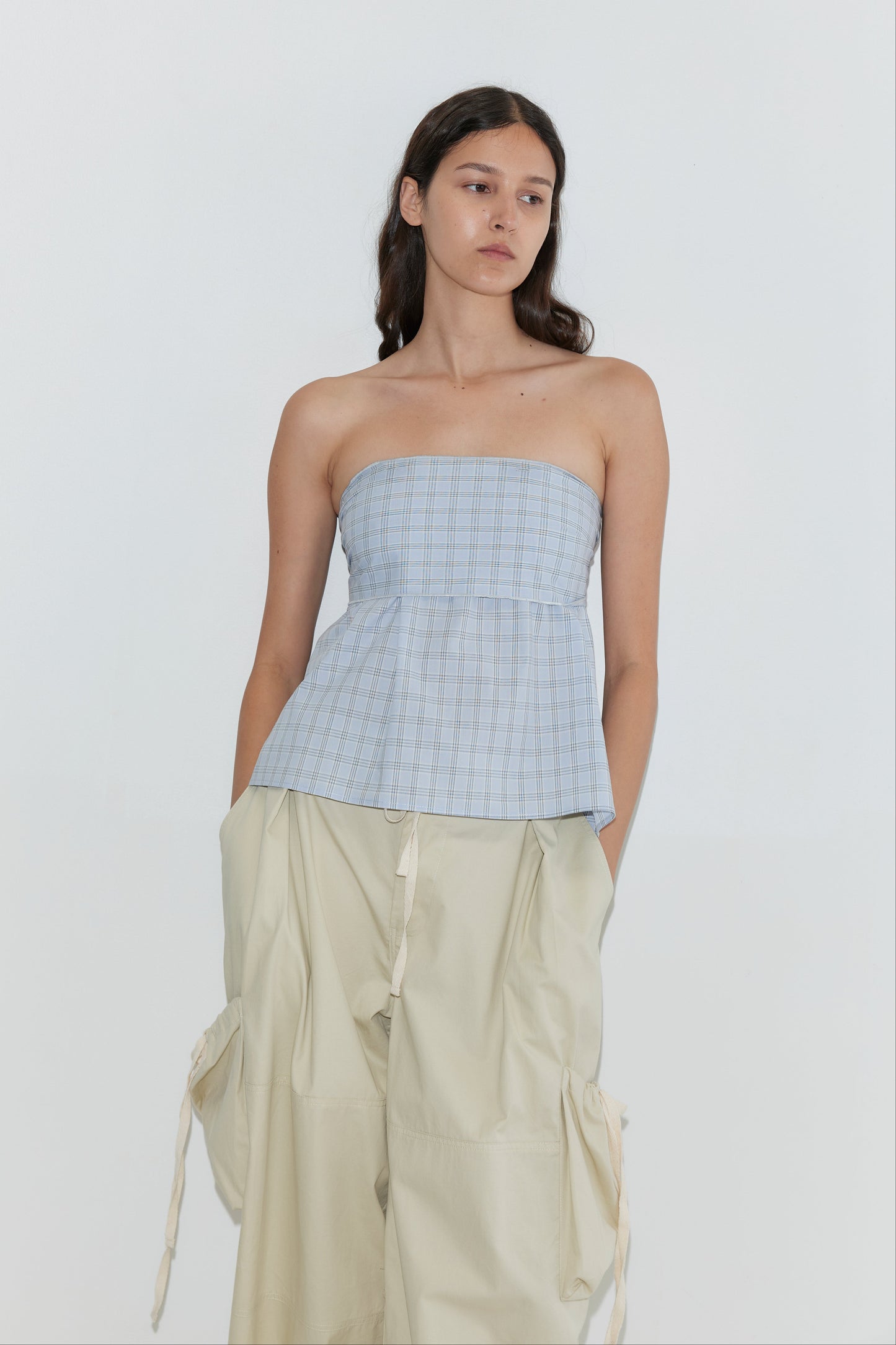 A woman with long brown hair wears a light blue strapless plaid top and Deiji Studios Cargo Pant - Stone featuring wide legs and drawstring details, standing against a plain white background with her hands in her pockets.